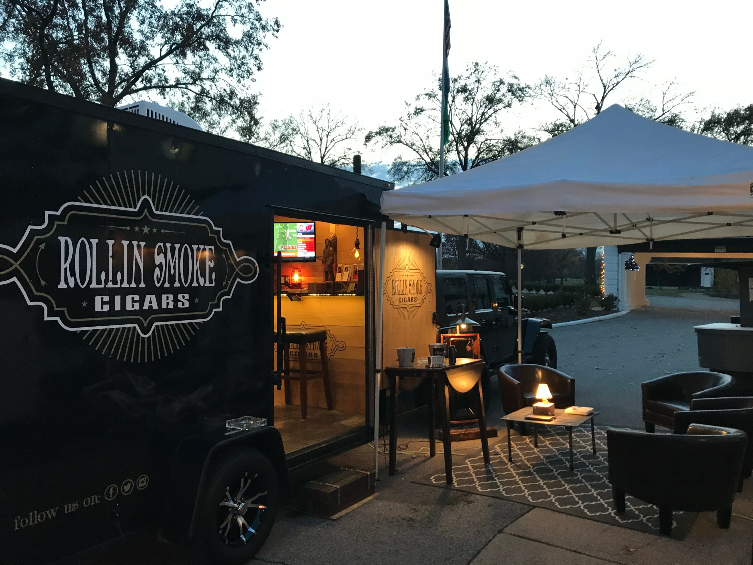 Outdoor setup with a white canopy, chairs, and a small table with a lamp, in front of a cigar shop sign.