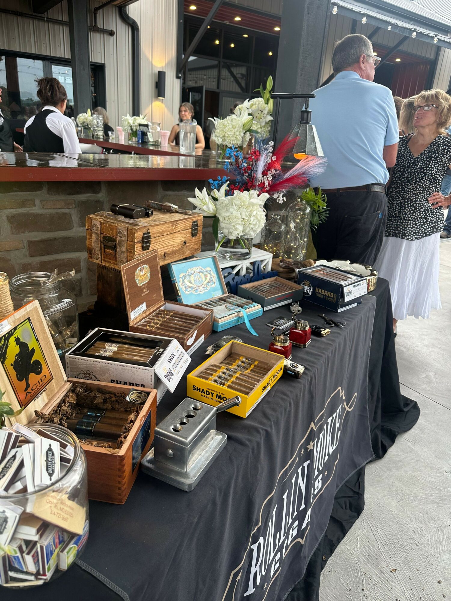 Table with various vintage items and books, people in background at an indoor event, flowers and decorations visible.