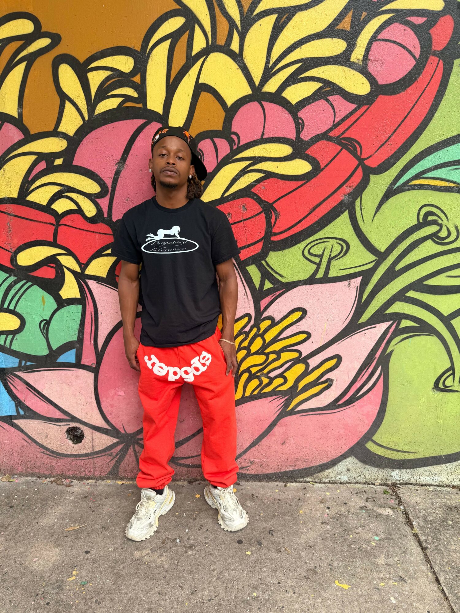 Person standing in front of colorful graffiti wall, wearing black shirt and red pants, on sidewalk.