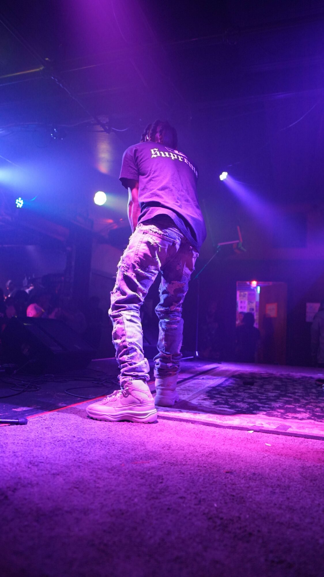 Person standing on stage with purple and blue lighting, facing away from the camera, in a performance setting.