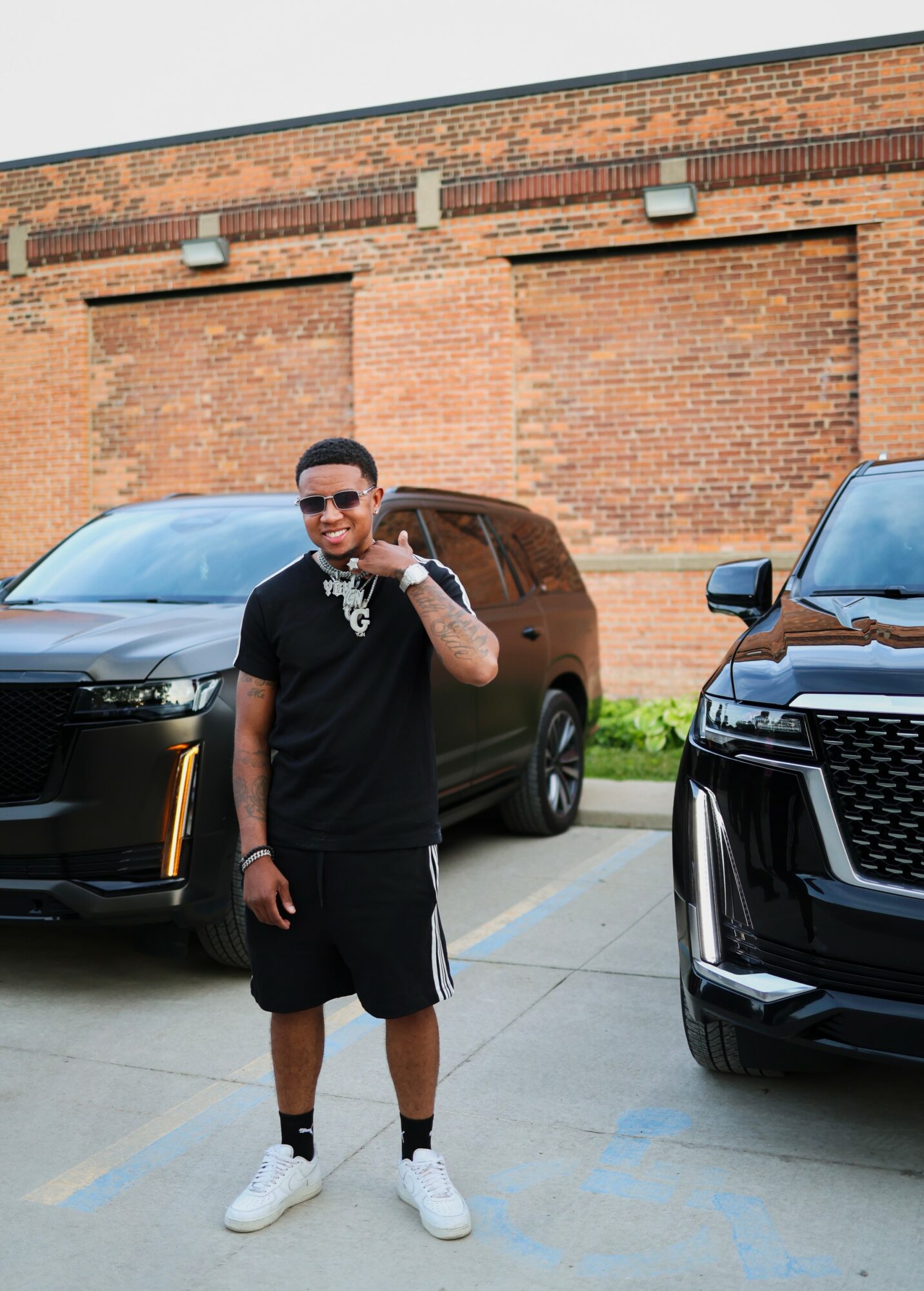 Young man standing between two black SUVs in front of a brick wall, wearing black clothing and white sneakers.