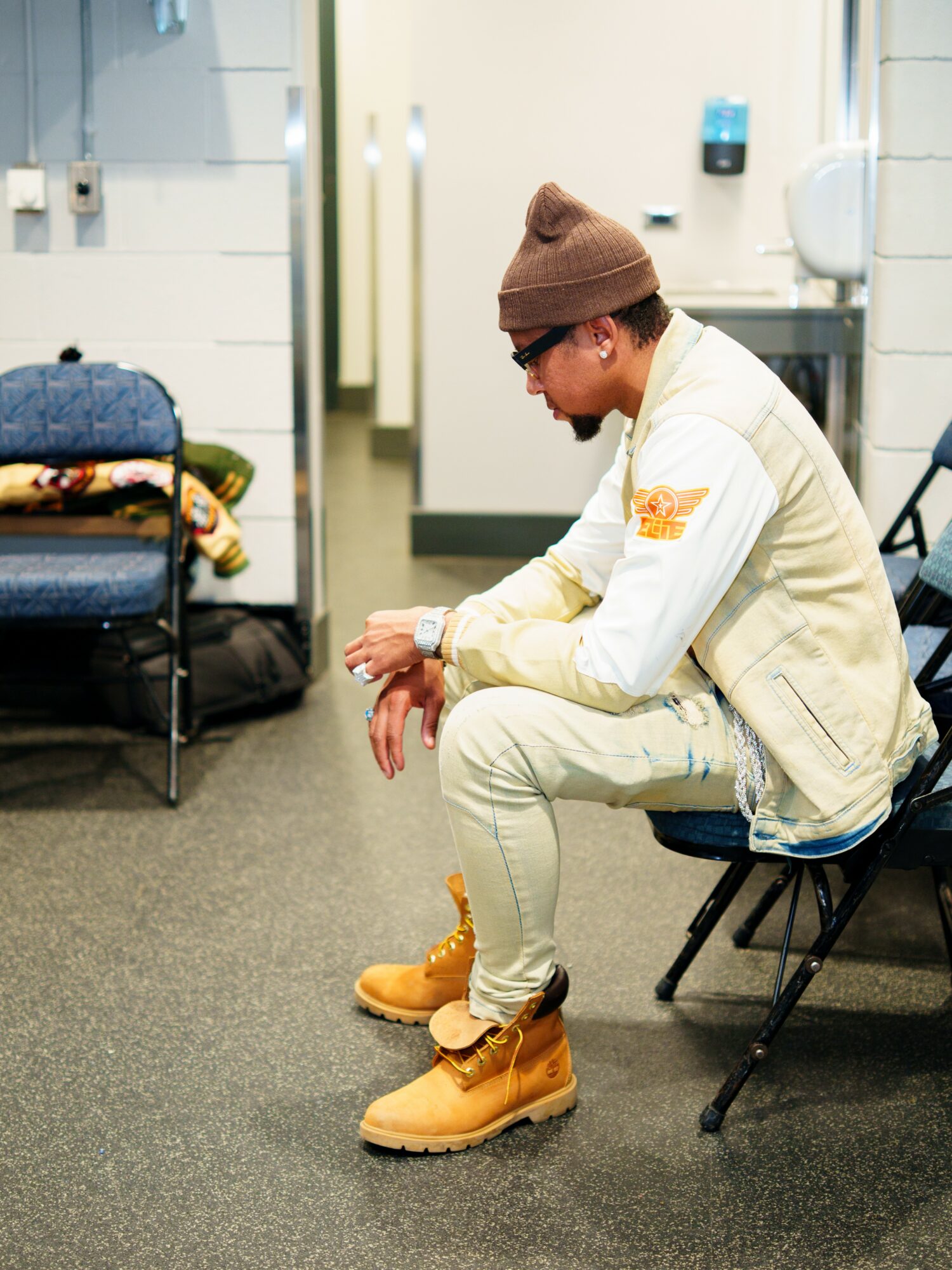 Person sitting on a chair in a room, adjusting their shoe, wearing a beanie, glasses, and beige clothing.