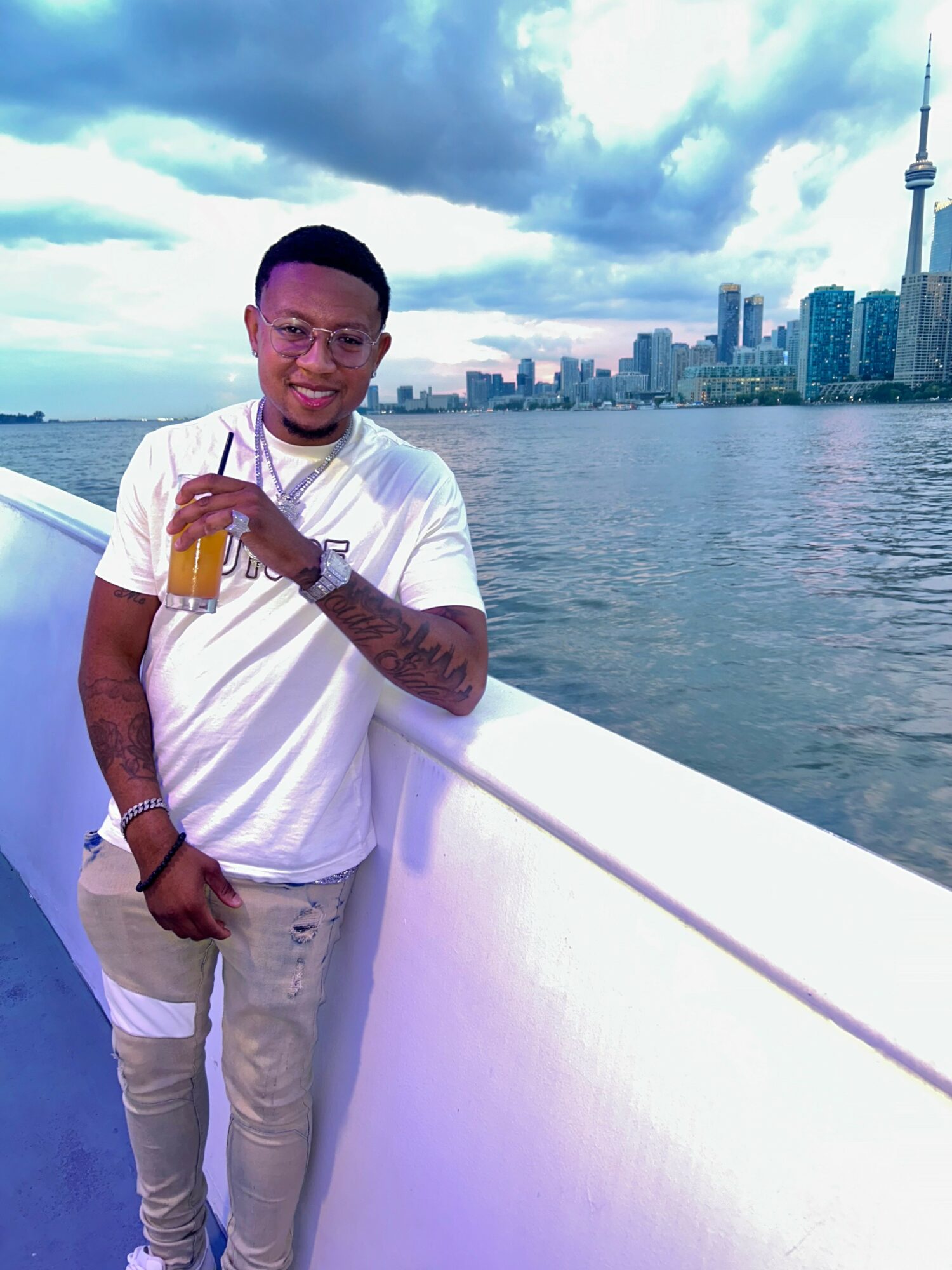 Young man with glasses and tattoos holding a drink, standing by a river with city skyline and CN Tower in background.