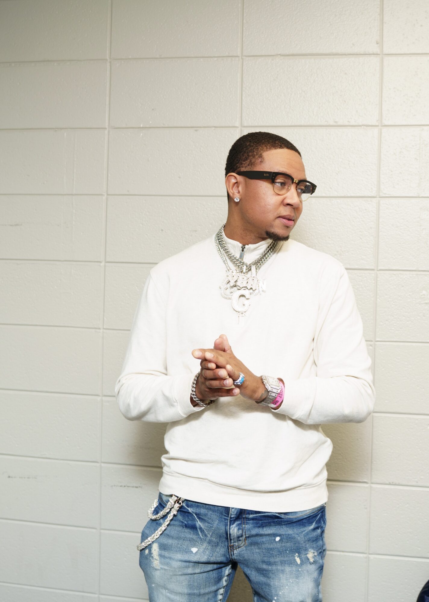 Young man with glasses and jewelry, wearing a white sweater and jeans, standing against a tiled wall, looking to the side.