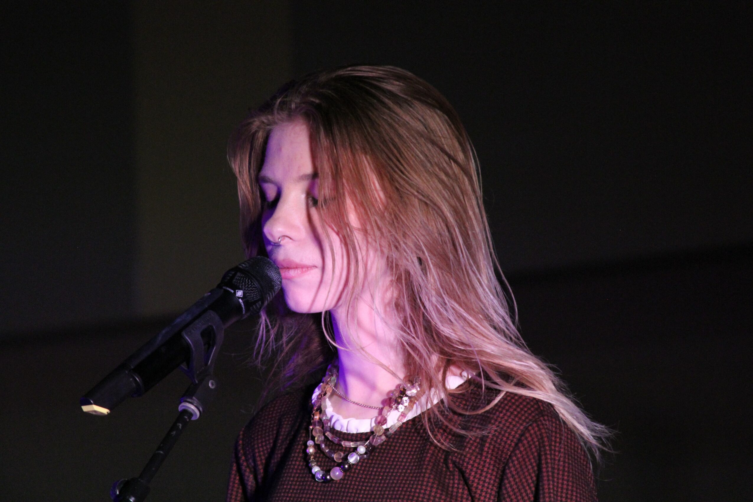 Woman with long hair wearing layered necklaces, singing into a microphone, with dark background.