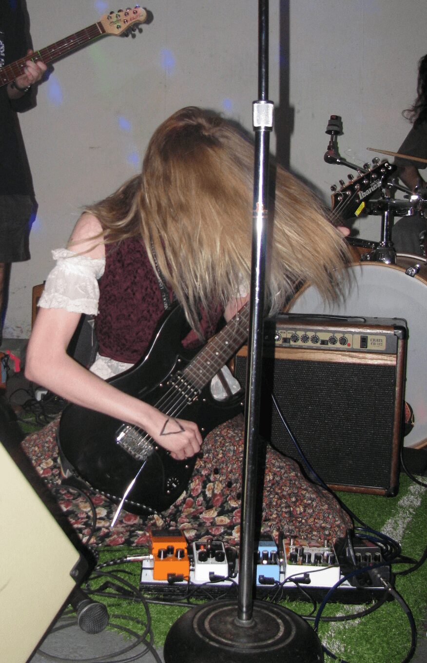Person with long hair playing a black electric guitar, surrounded by musical equipment and pedals, in a room.