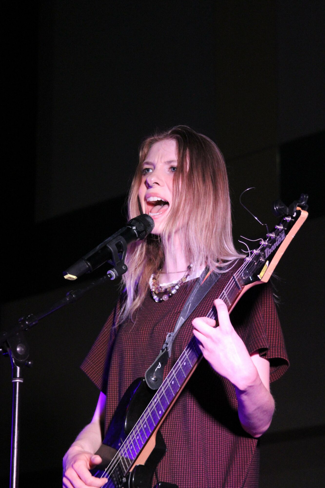Woman singing into a microphone while playing an acoustic guitar on stage, with dark background.