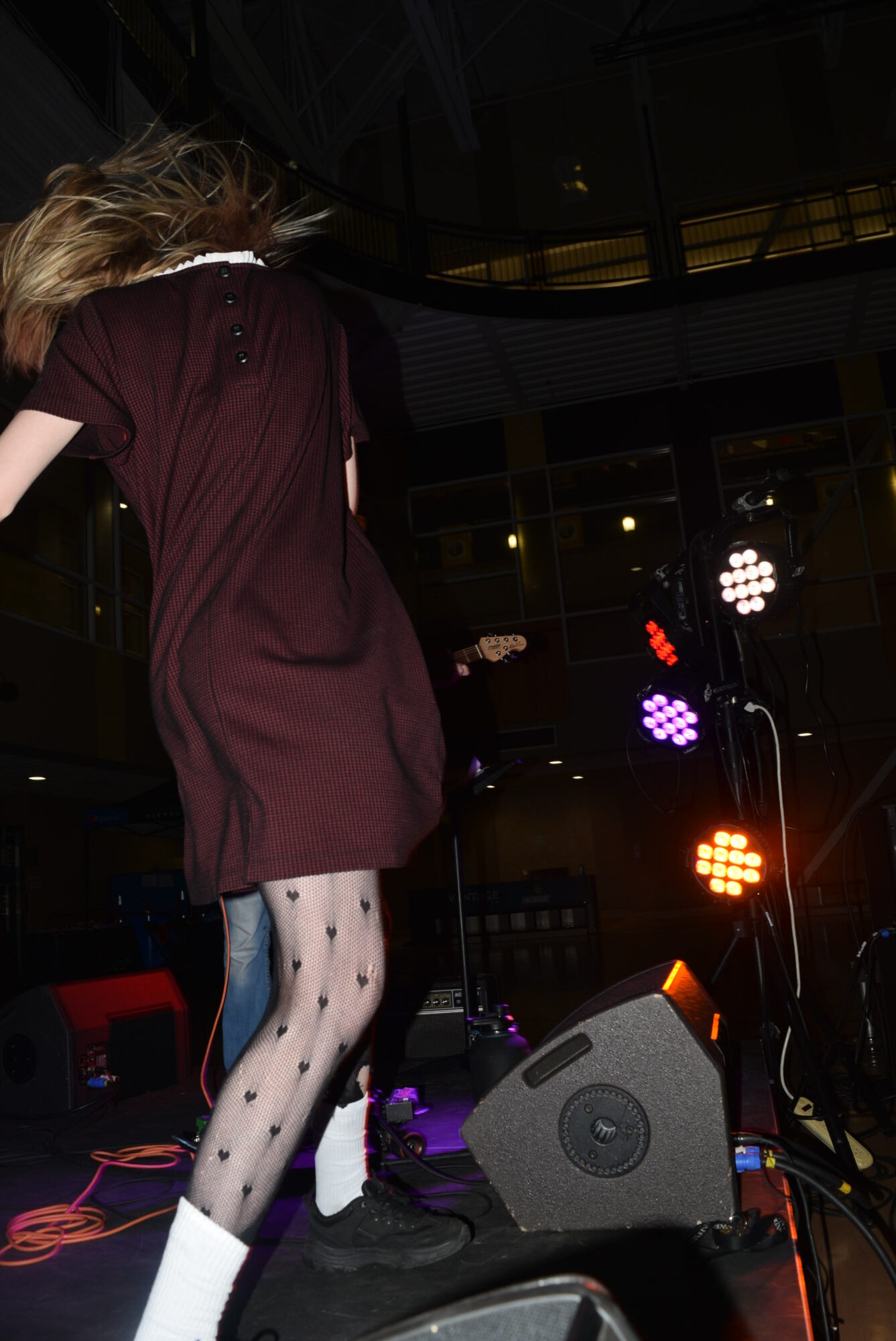 Person with long hair wearing a dark dress and polka dot tights standing on stage with colorful lights in background.