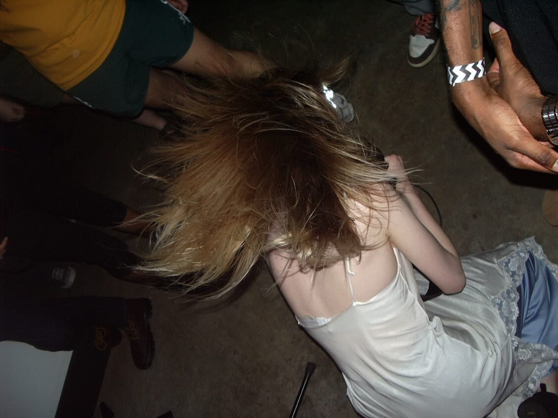 Person with long, blonde hair sitting on the floor, wearing a white dress, surrounded by other people.