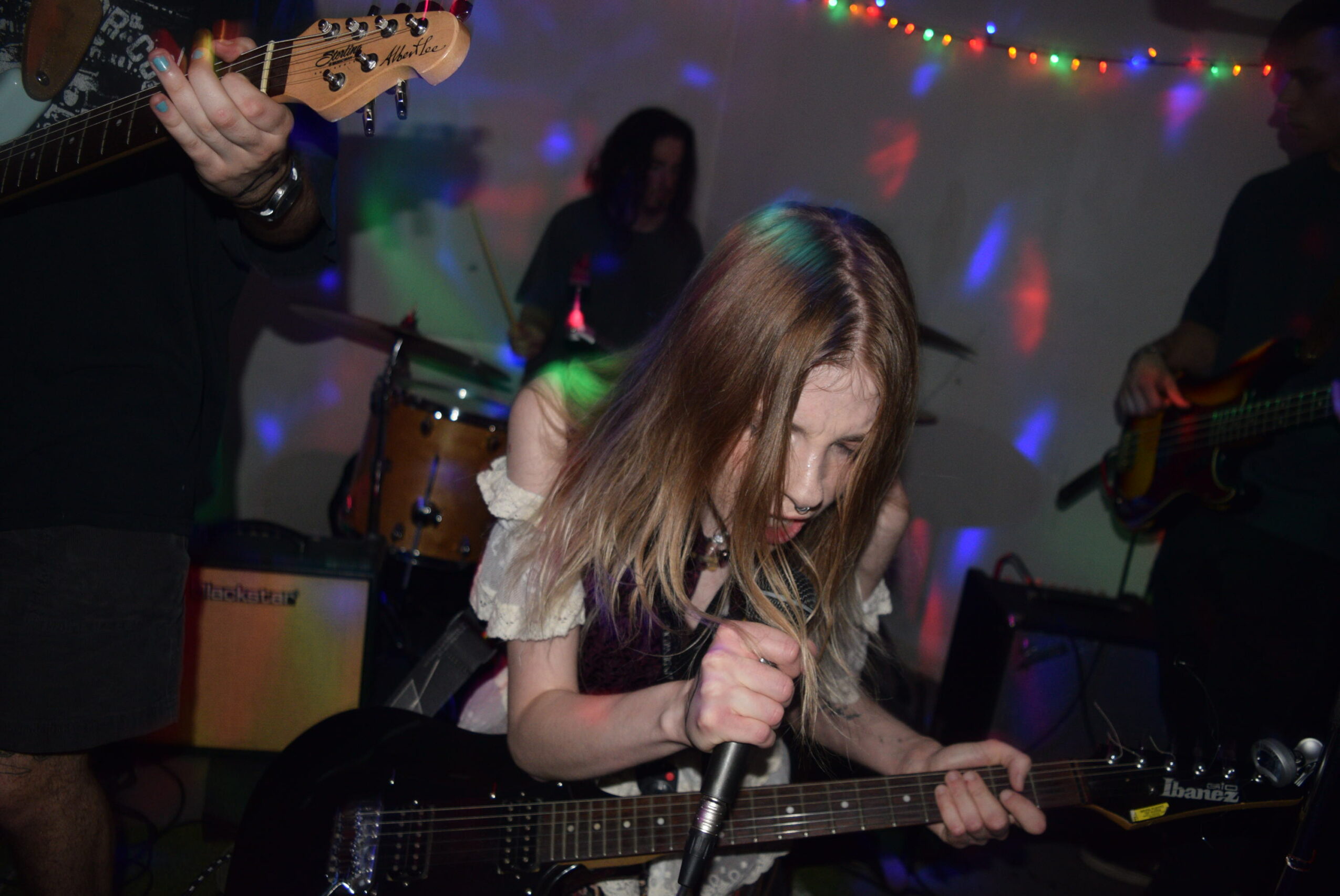 Young woman with long hair sings into a microphone while playing an electric guitar at a party with colorful lights.