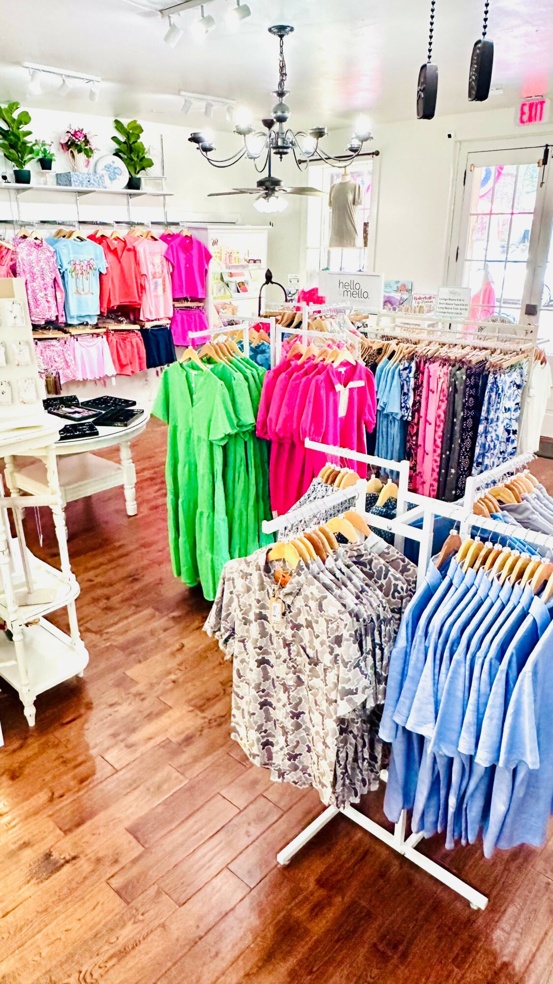 Clothing store interior with colorful dresses on racks, shelves, and a chandelier, wooden floor, and large window.