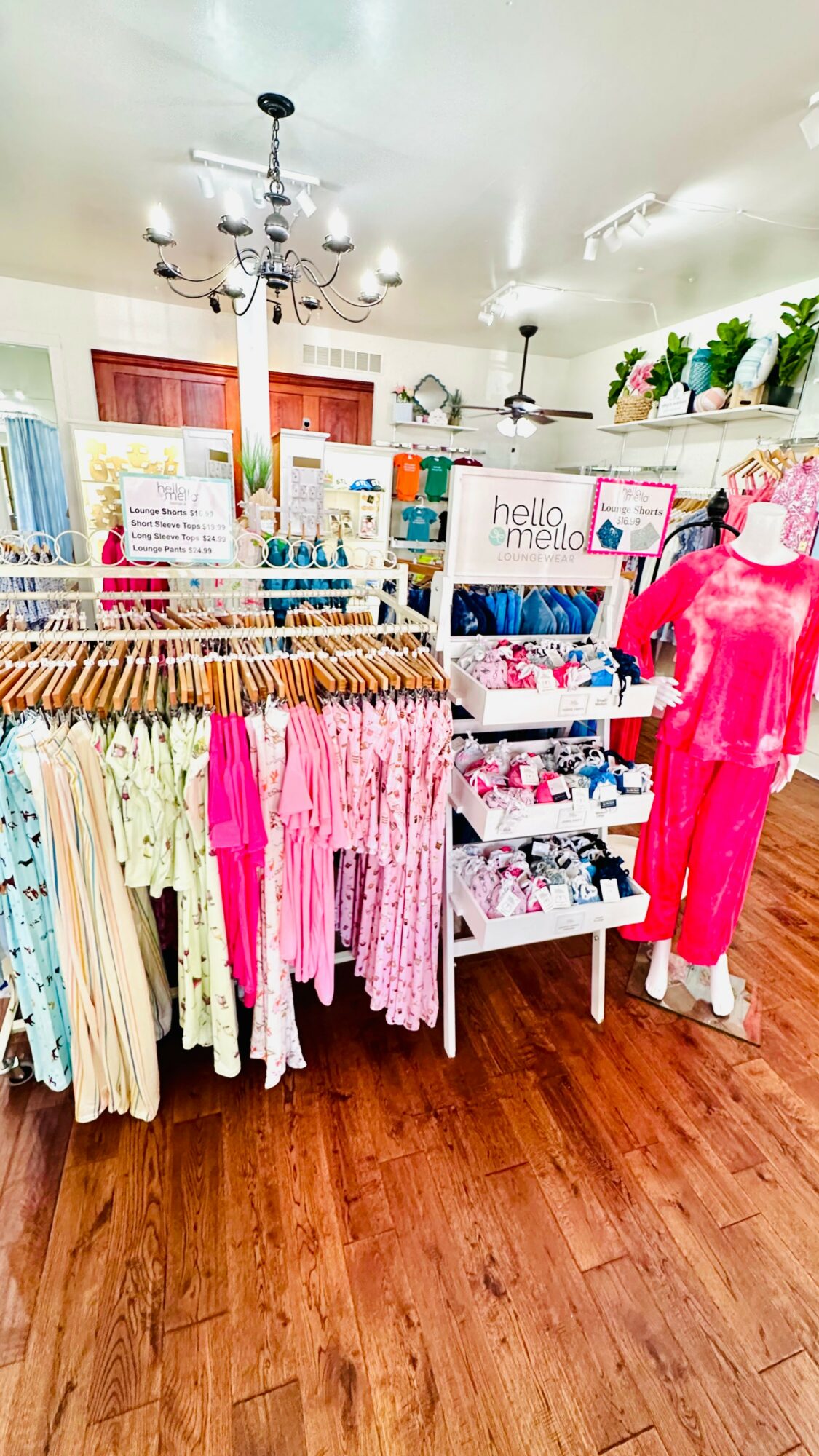 Clothing display with pink and white garments, shelves with books, and potted plants on top, in a store interior.