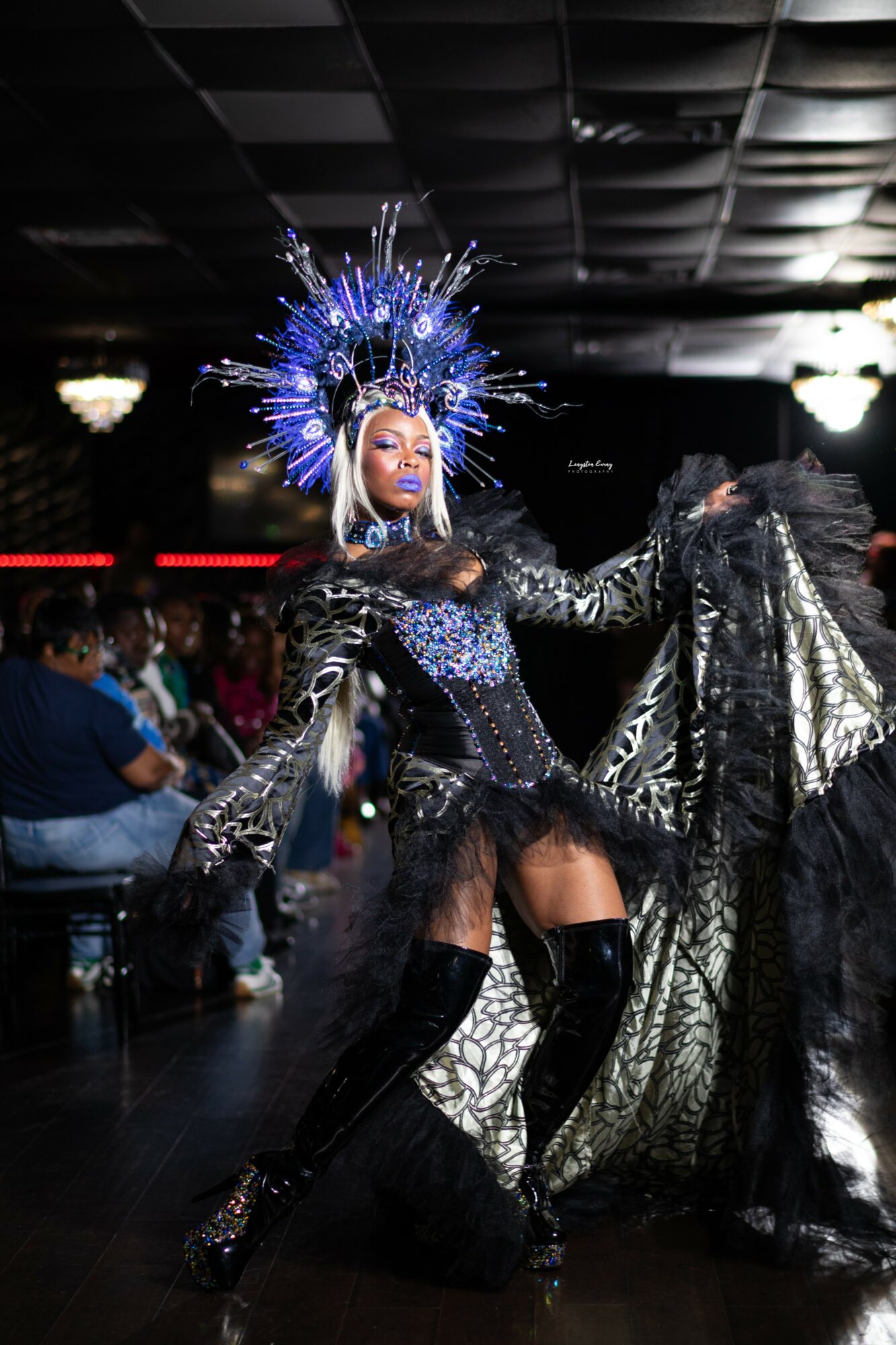 Person in elaborate costume with a blue headdress, black lace dress, and thigh-high boots performing on stage.