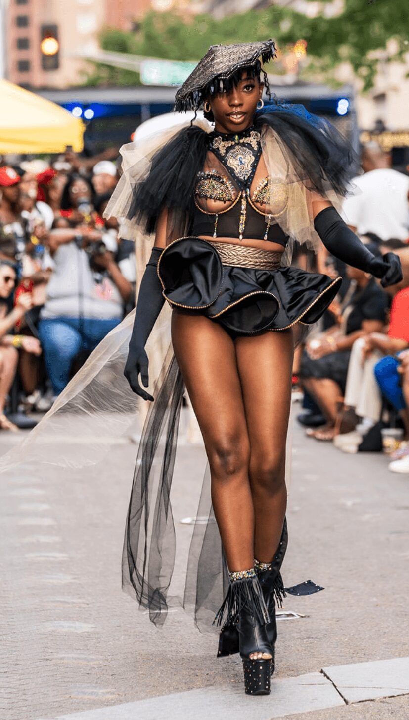 Model walking on runway in elaborate outfit with black platform shoes, hat, and sheer fabric, audience watching in background.