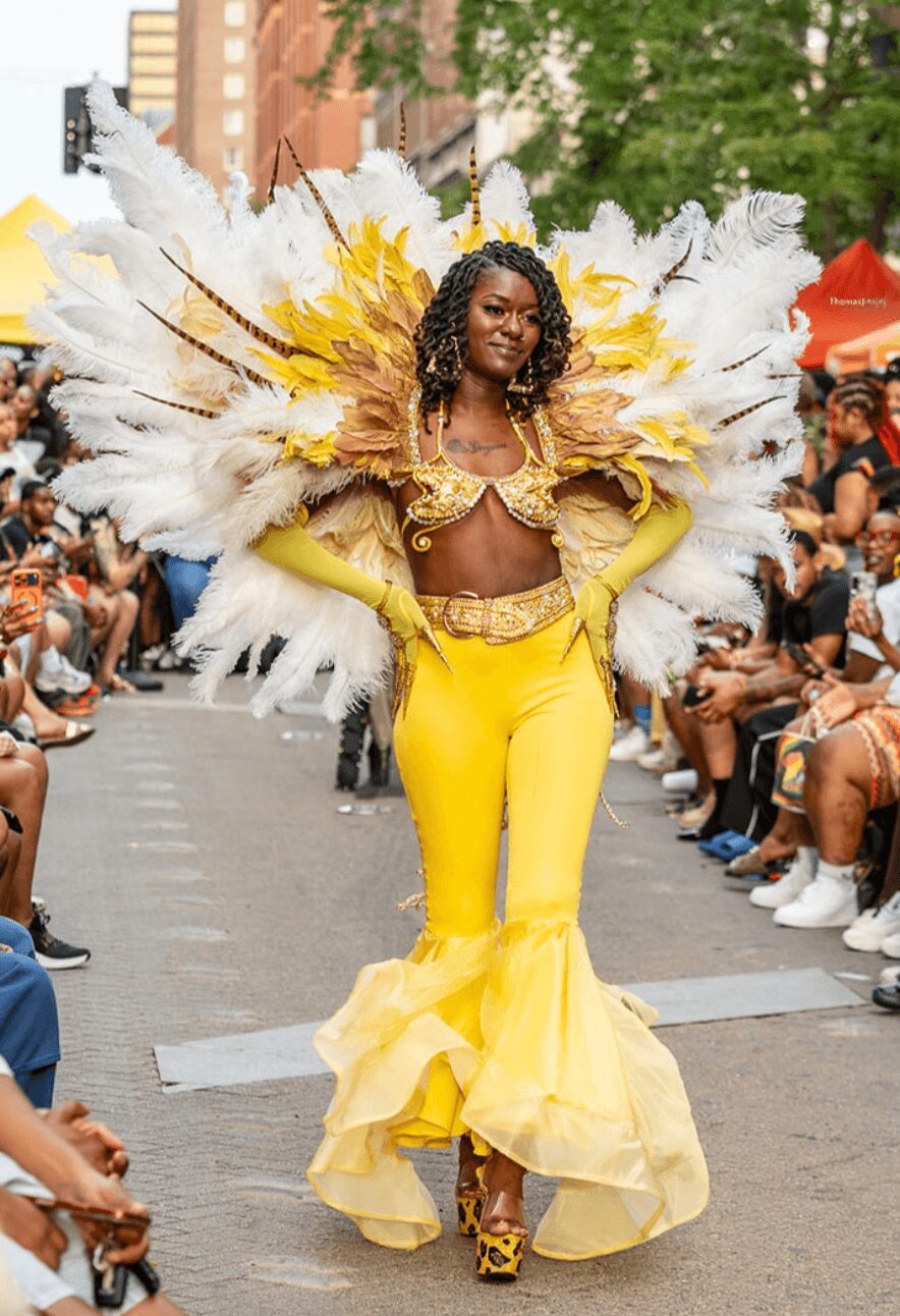 Woman in yellow costume with large feathered wings walking in parade, surrounded by seated spectators.