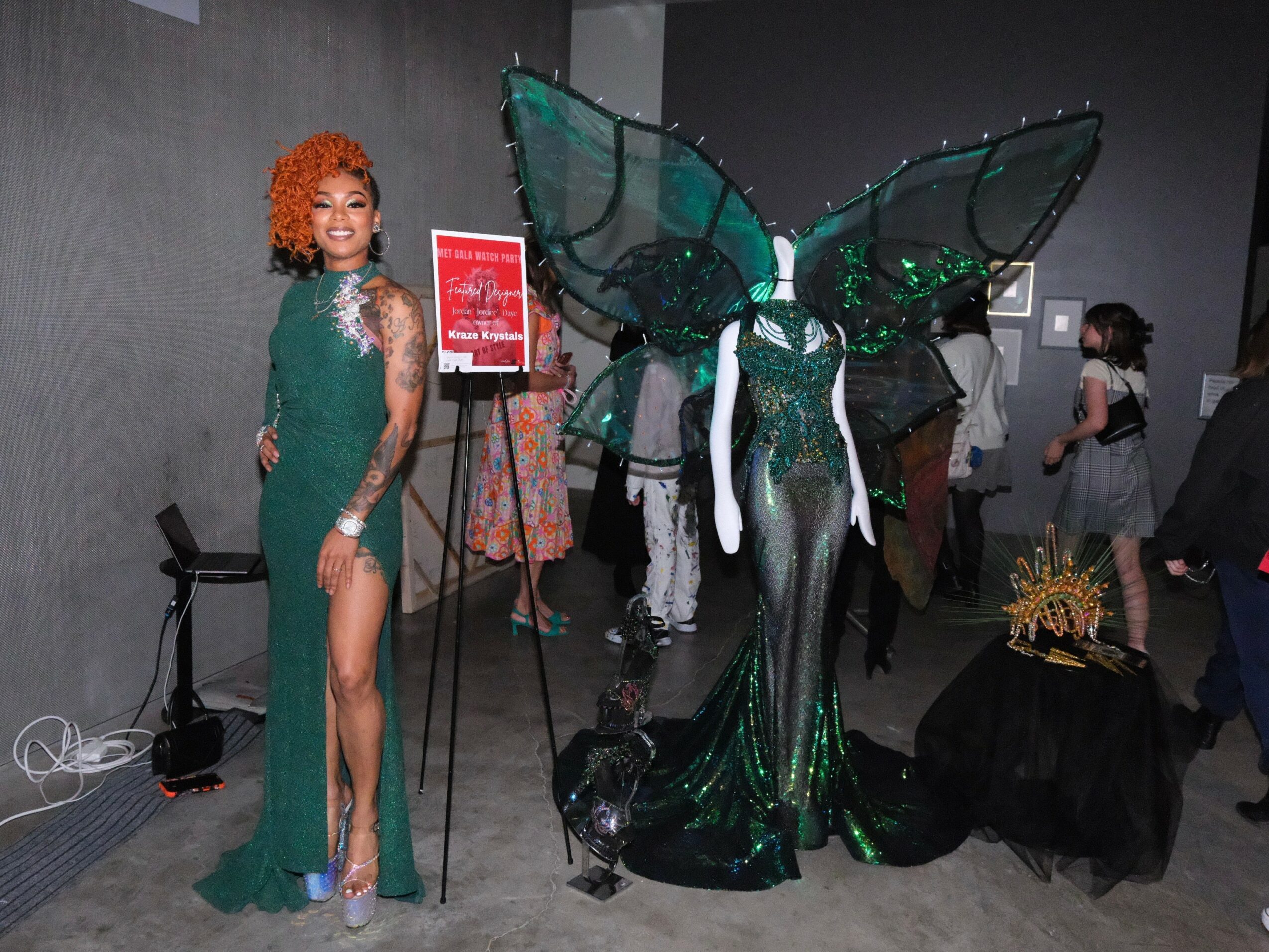 Woman in a green dress with a high slit, standing next to a large butterfly sculpture with green and black wings.