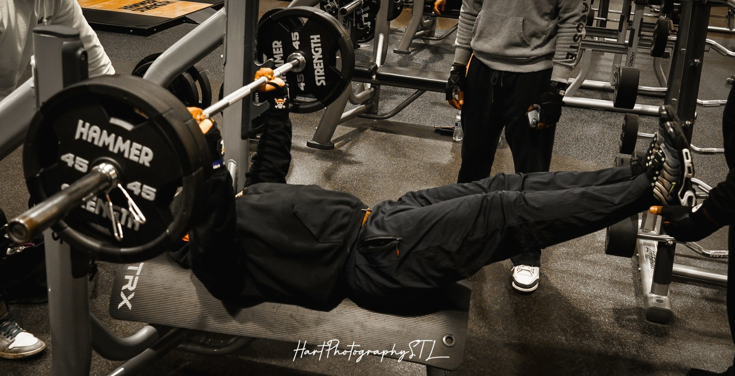 Person lying on a bench lifting a barbell in a gym, with people standing nearby.
