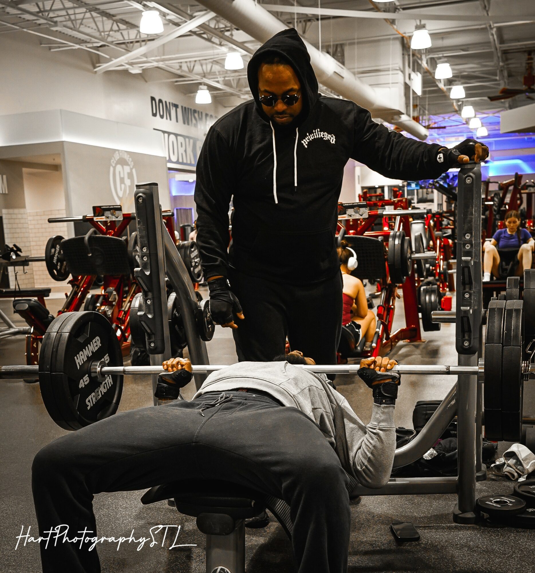 Person lying on bench press with barbell, trainer spotting in gym with weightlifting equipment in background.