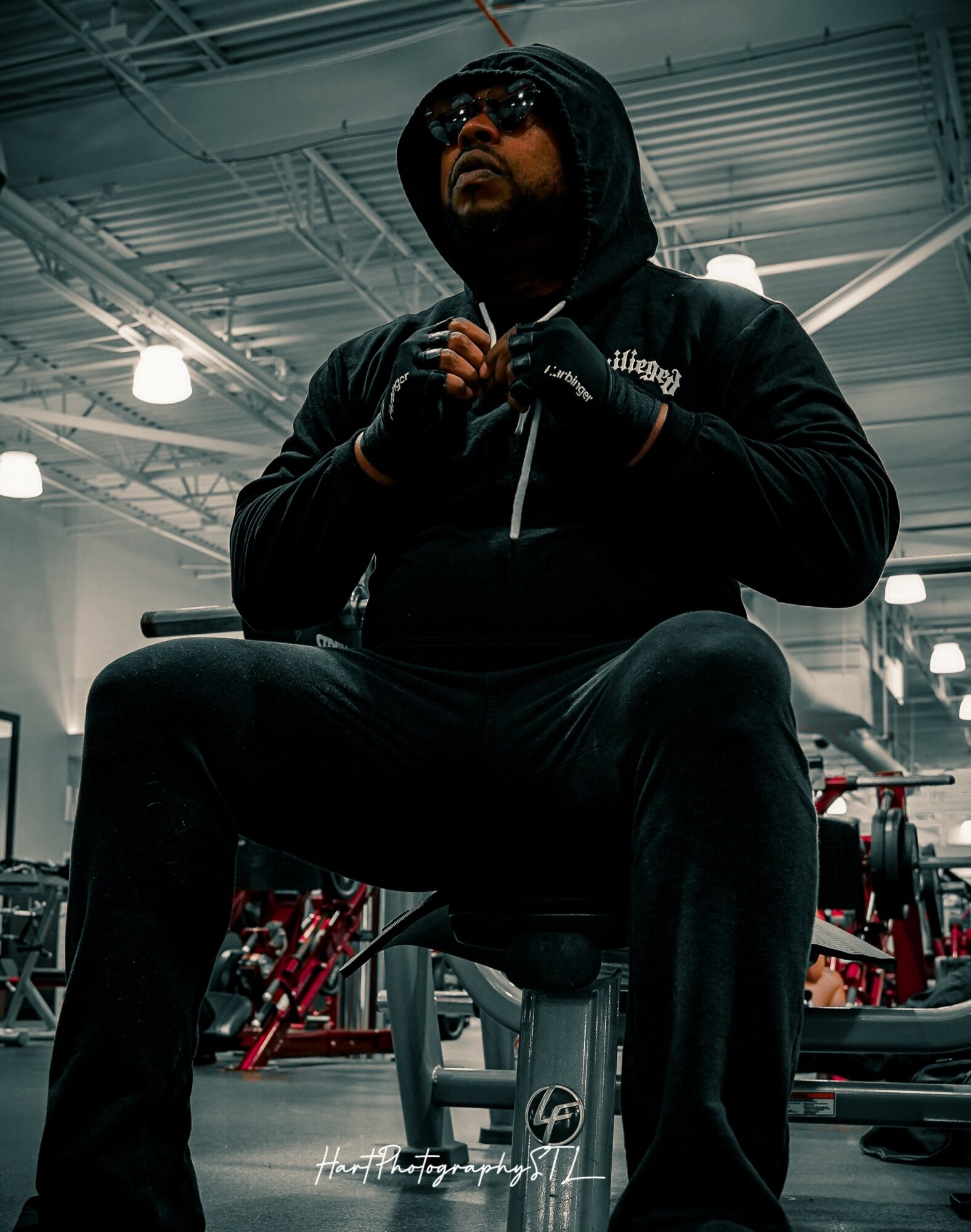 Person sitting on a gym bench, wearing sunglasses and a hoodie, with gym equipment in the background.