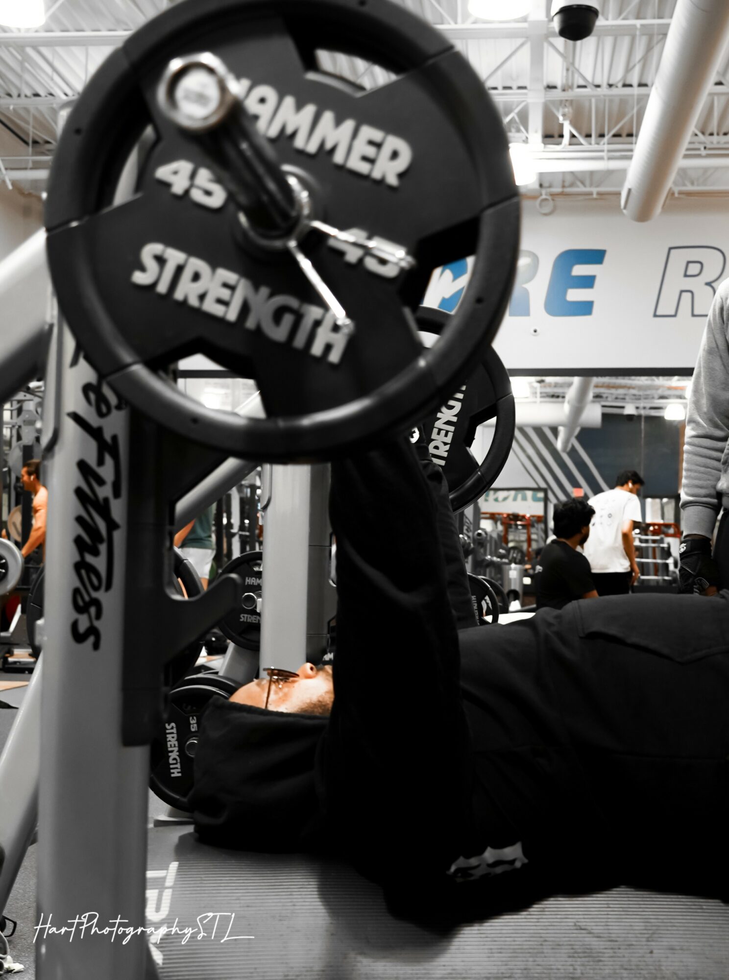 Person lying on the floor lifting a weight barbell in a gym, with gym equipment and people in the background.
