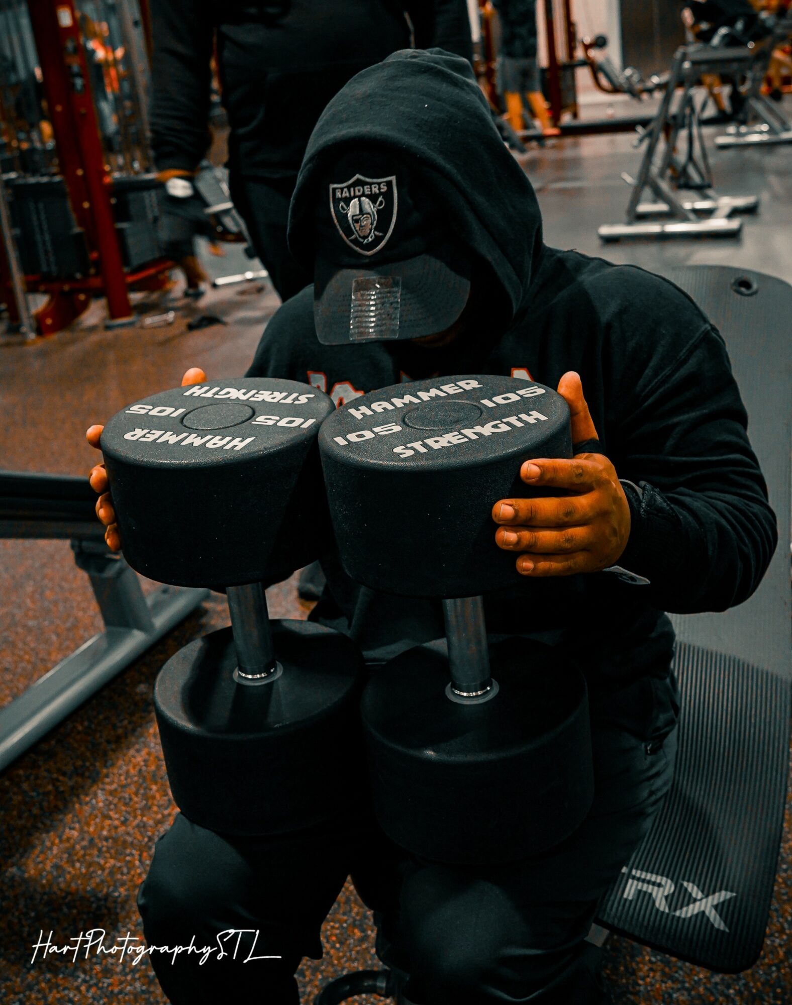 Person in black hoodie with hood up, holding two large dumbbells in a gym, with gym equipment in background.