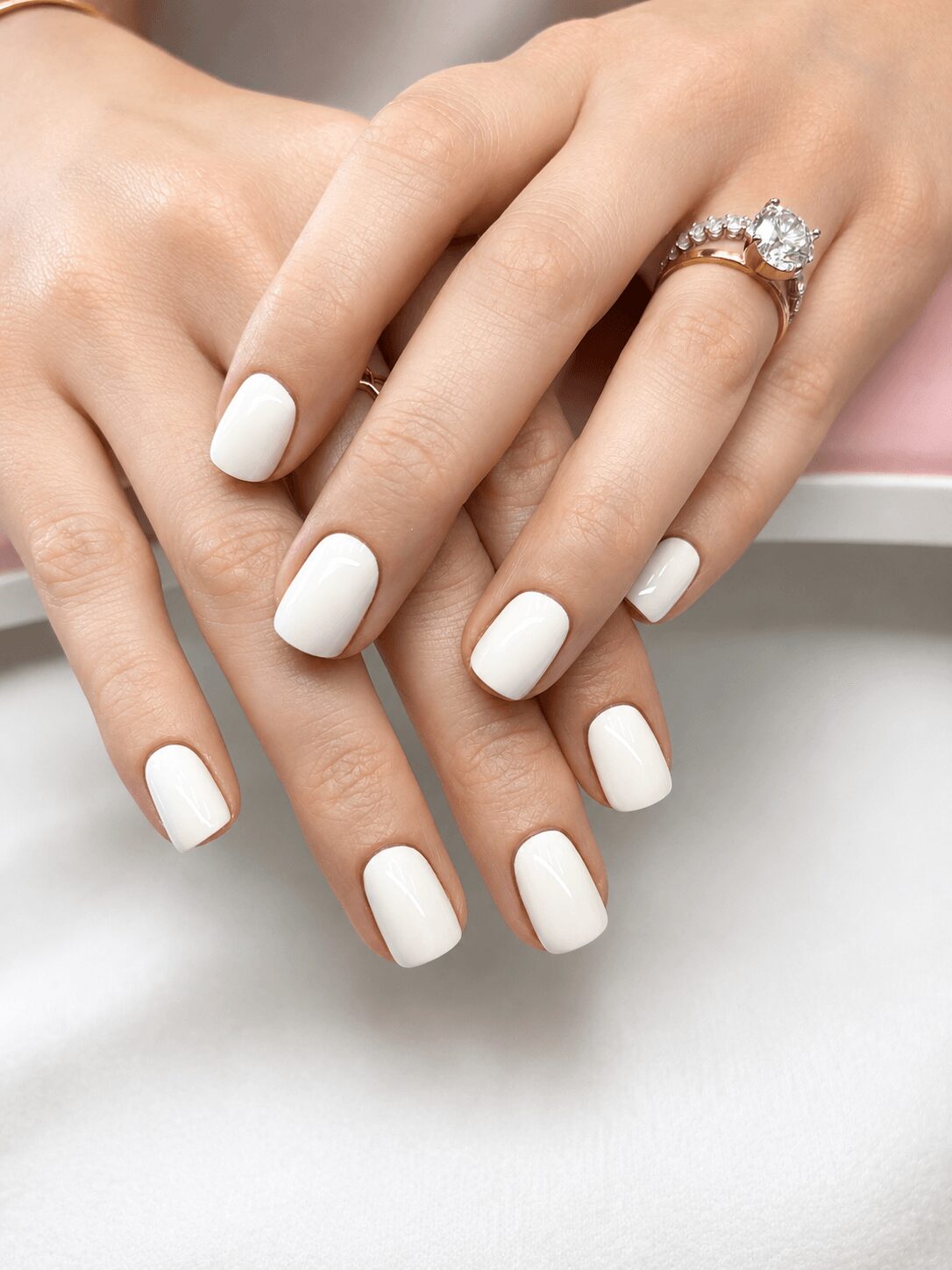 Two hands with white manicured nails, one hand wearing a ring with a large gemstone, resting on a white surface.