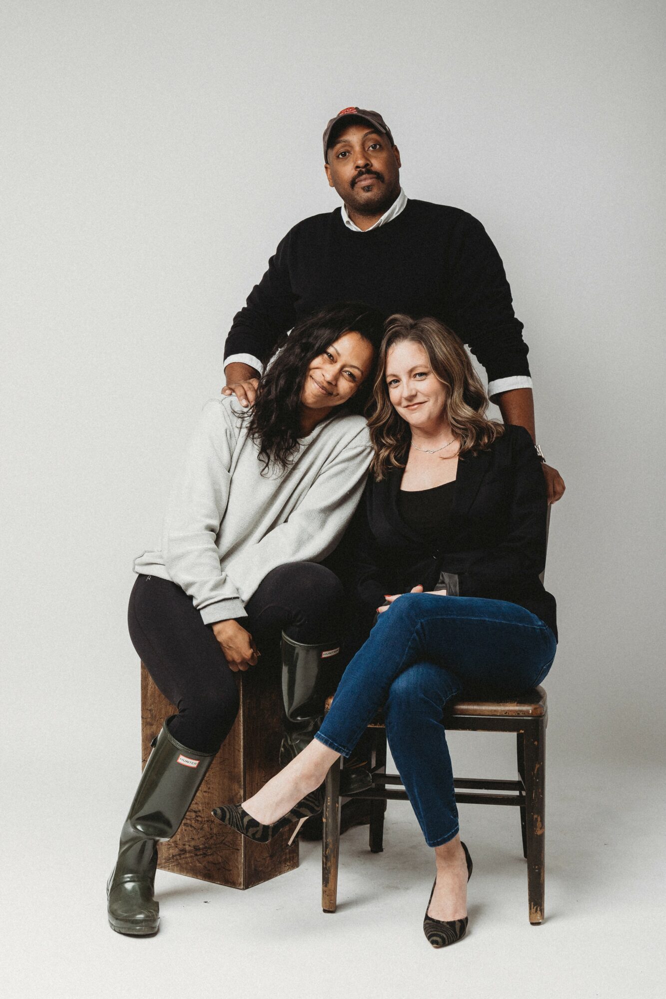Three people, two women sitting on chairs and one man standing behind them, in a studio with a plain background.