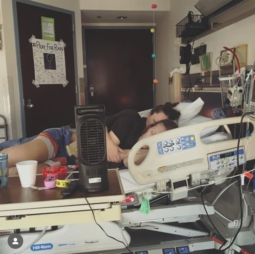 Person lying in hospital bed with medical equipment, fan, and colorful decorations in the room.