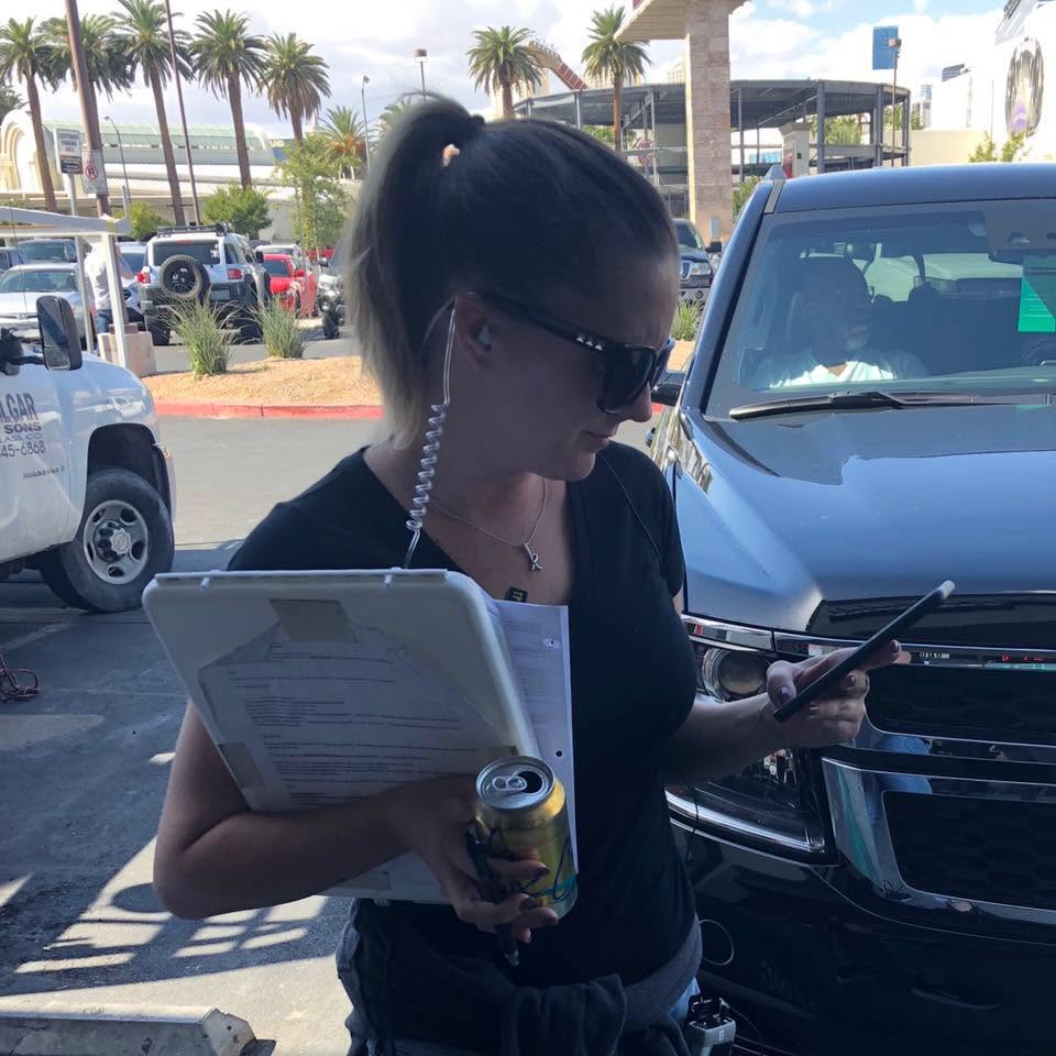 Woman with ponytail and sunglasses holding a tablet, a can, and a phone outdoors near parked cars.