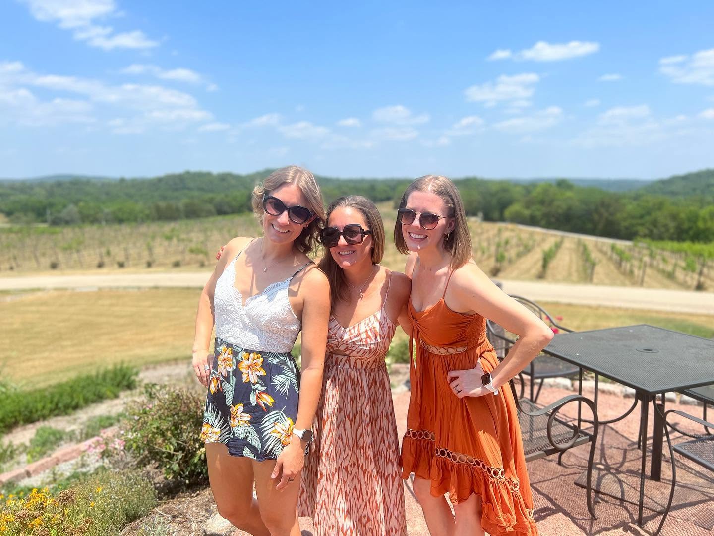 Three women standing outdoors on a sunny day, smiling, with a landscape of trees and sky in the background.