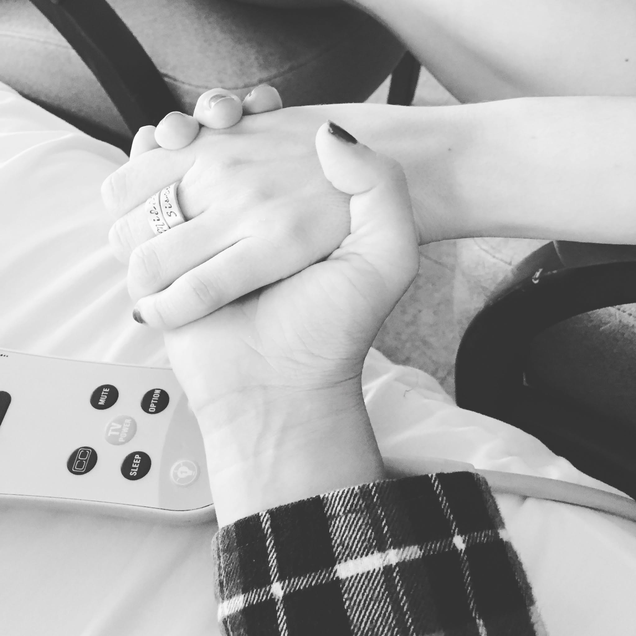 Two people hold hands, one with a ring, in a black-and-white photo. A remote control is visible nearby.
