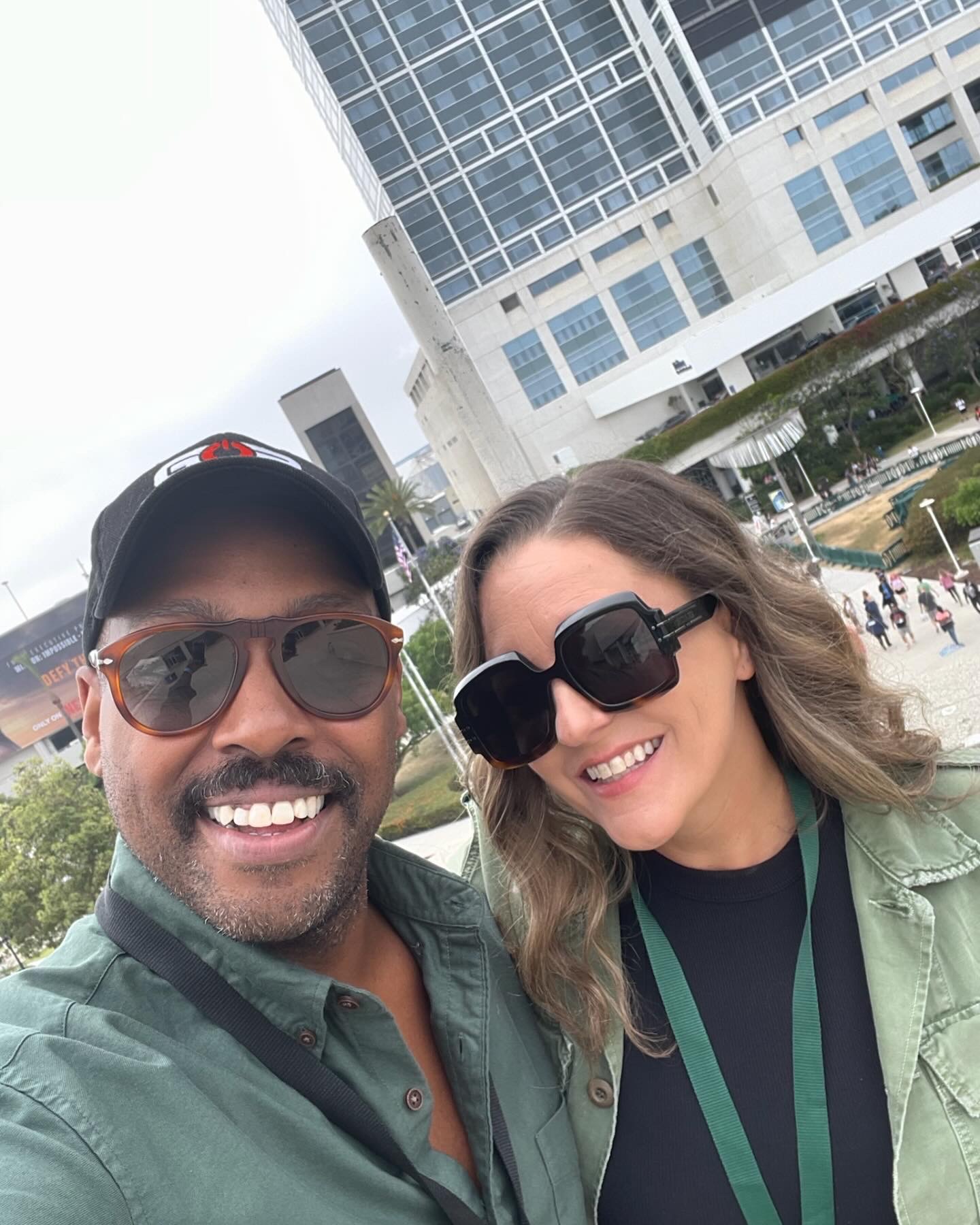 Two people smiling outdoors with a modern glass building in the background.
