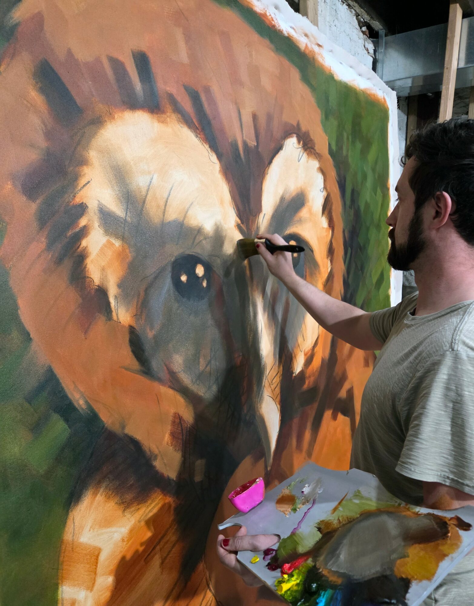Person painting a large, realistic bear portrait on a wall with a brush, holding a palette, in an indoor setting.