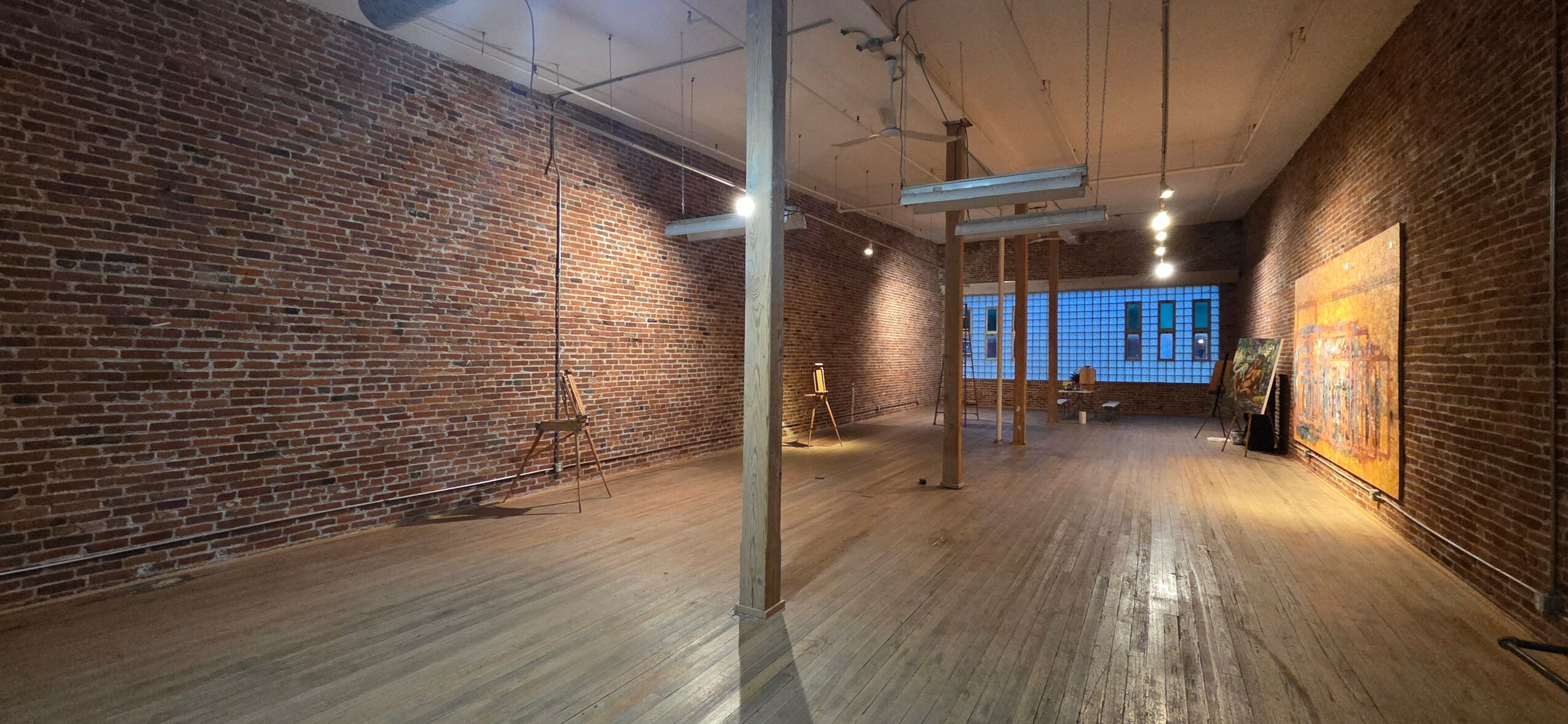 Empty art gallery with brick walls, wooden floor, and artwork on the right wall, illuminated by ceiling lights.