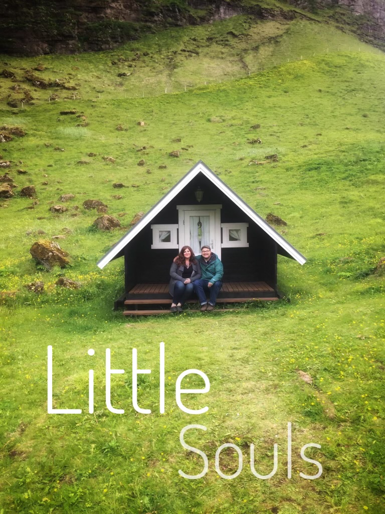Two people sit on a small porch in front of a house with a steep roof, surrounded by green grass and hillside.