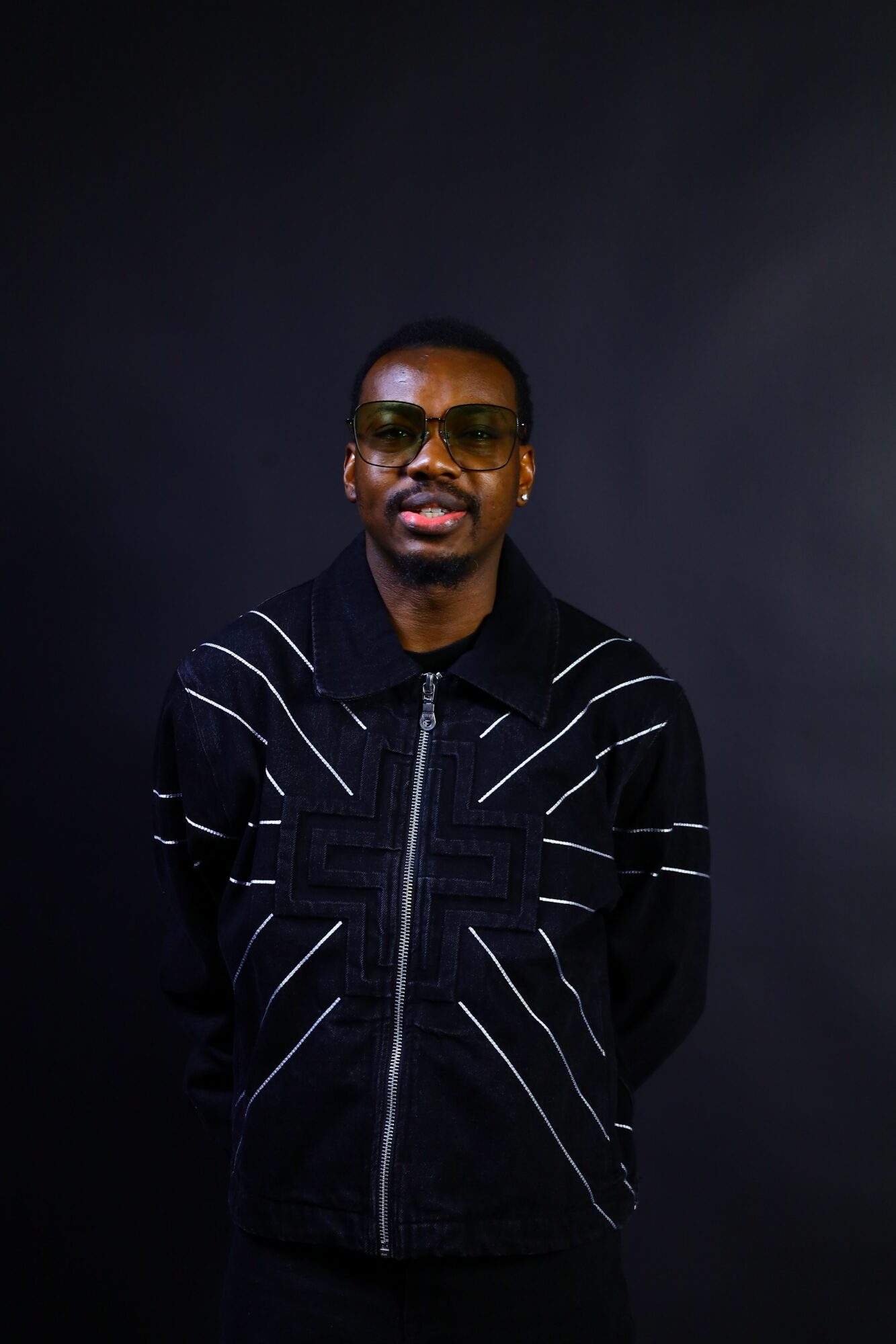 Person wearing sunglasses and a black jacket with white stripes, standing against a dark background.