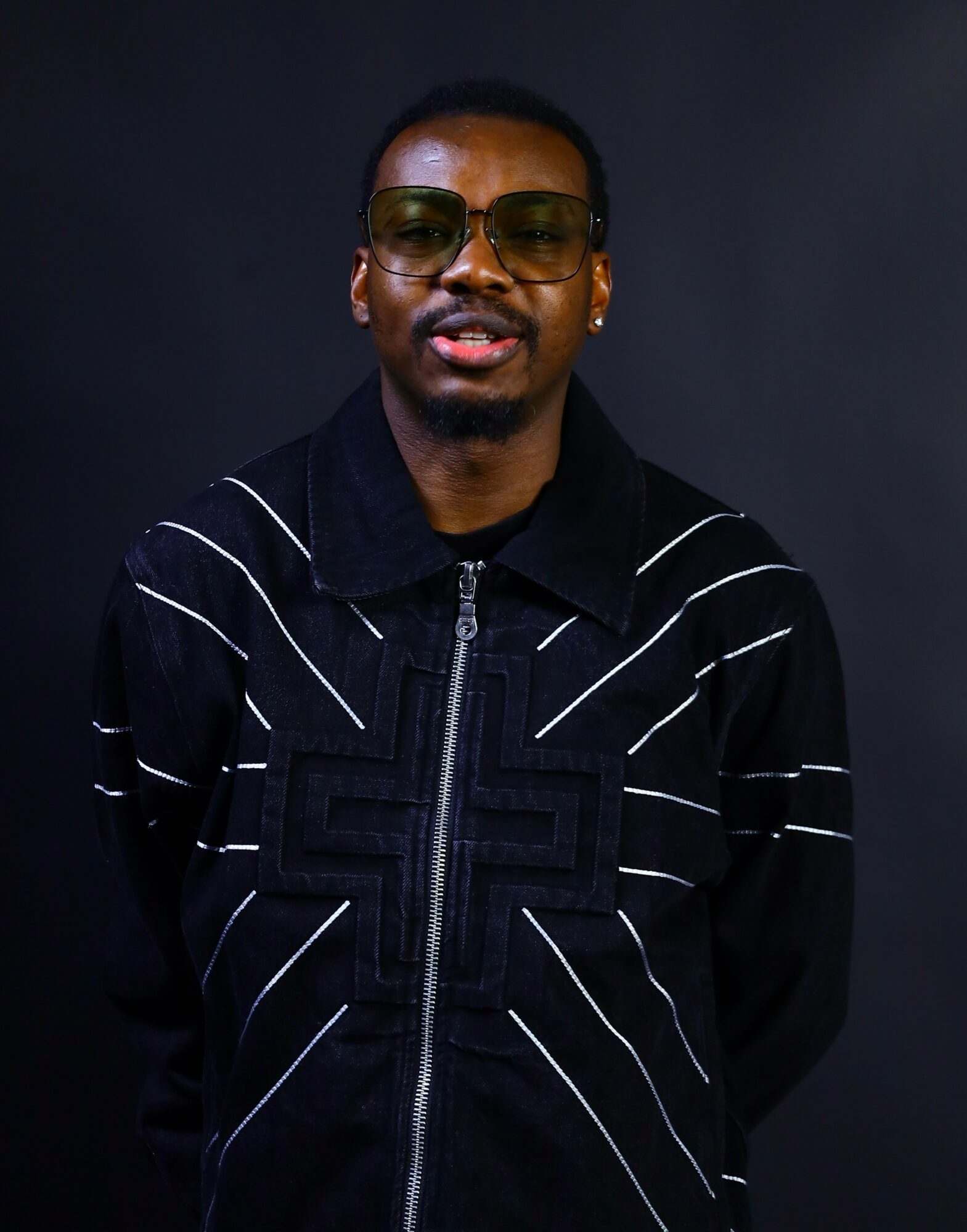 Young man wearing glasses and a black jacket with white lines, standing against a dark background.