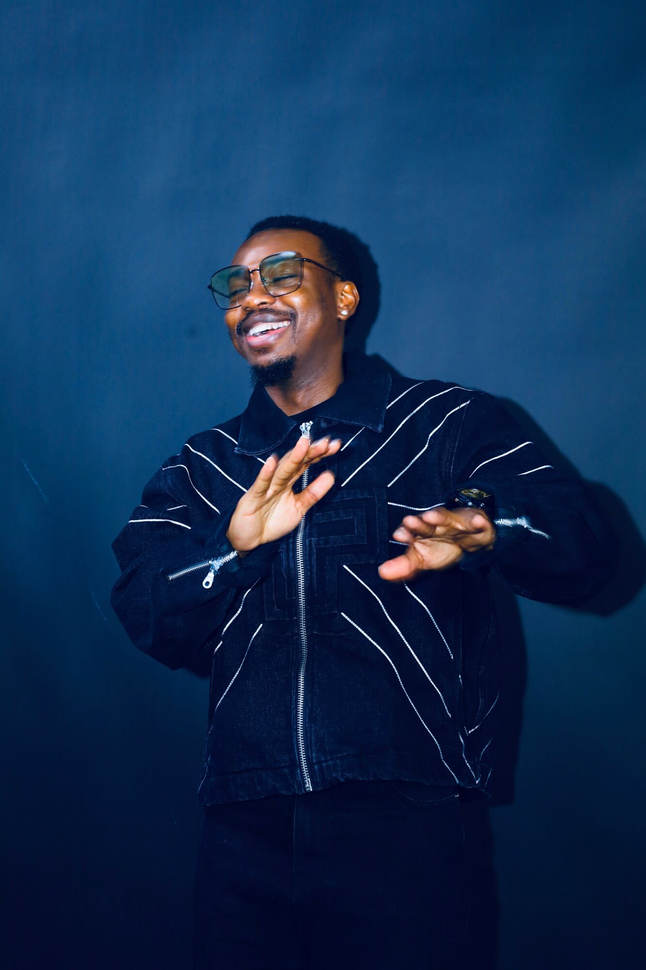 Person smiling, wearing glasses and a black jacket with white stripes, gesturing with hands against a dark blue background.