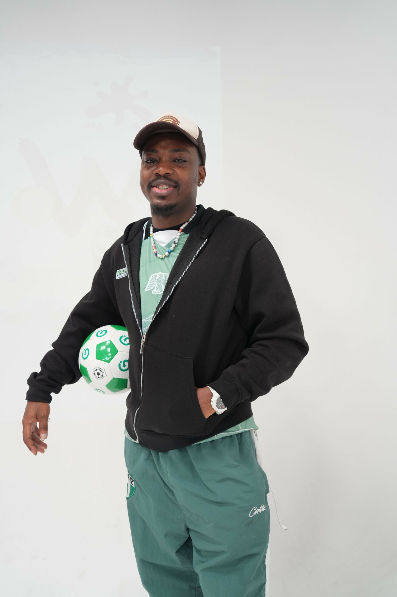 Person standing with a soccer ball, wearing a black jacket and green pants, against a plain white background.