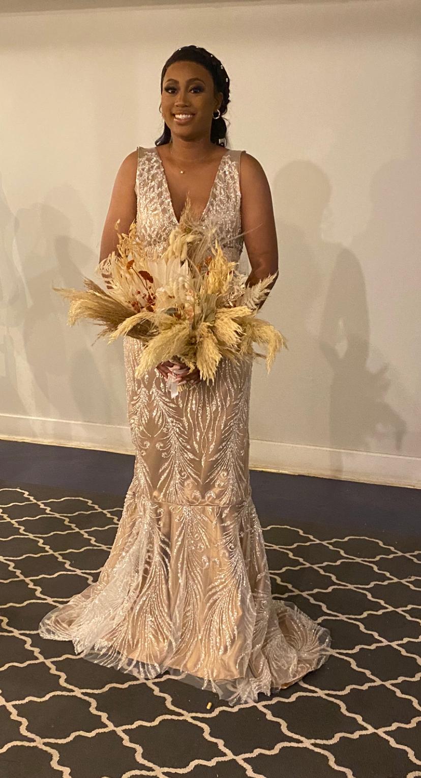 Woman in a detailed, elegant gown holding a large bouquet of dried flowers, standing on a patterned carpet.