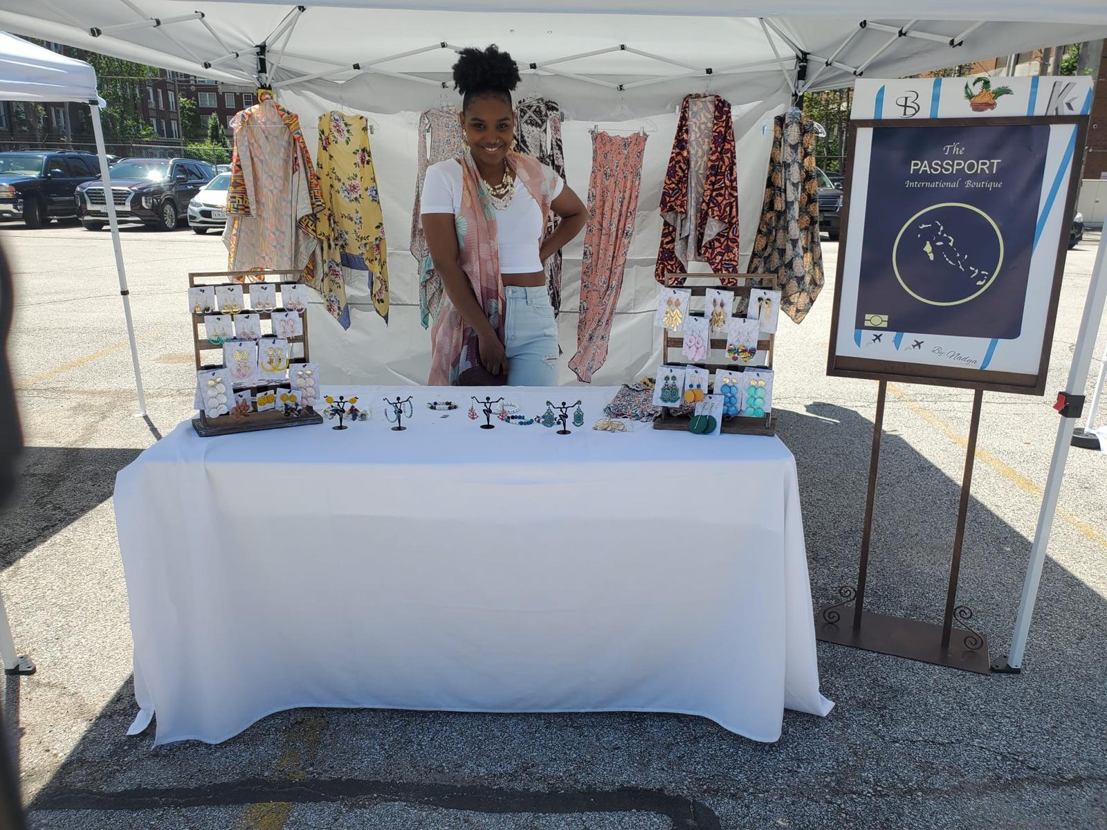 Woman standing behind a table with jewelry and clothing at an outdoor market stall, with a passport sign nearby.
