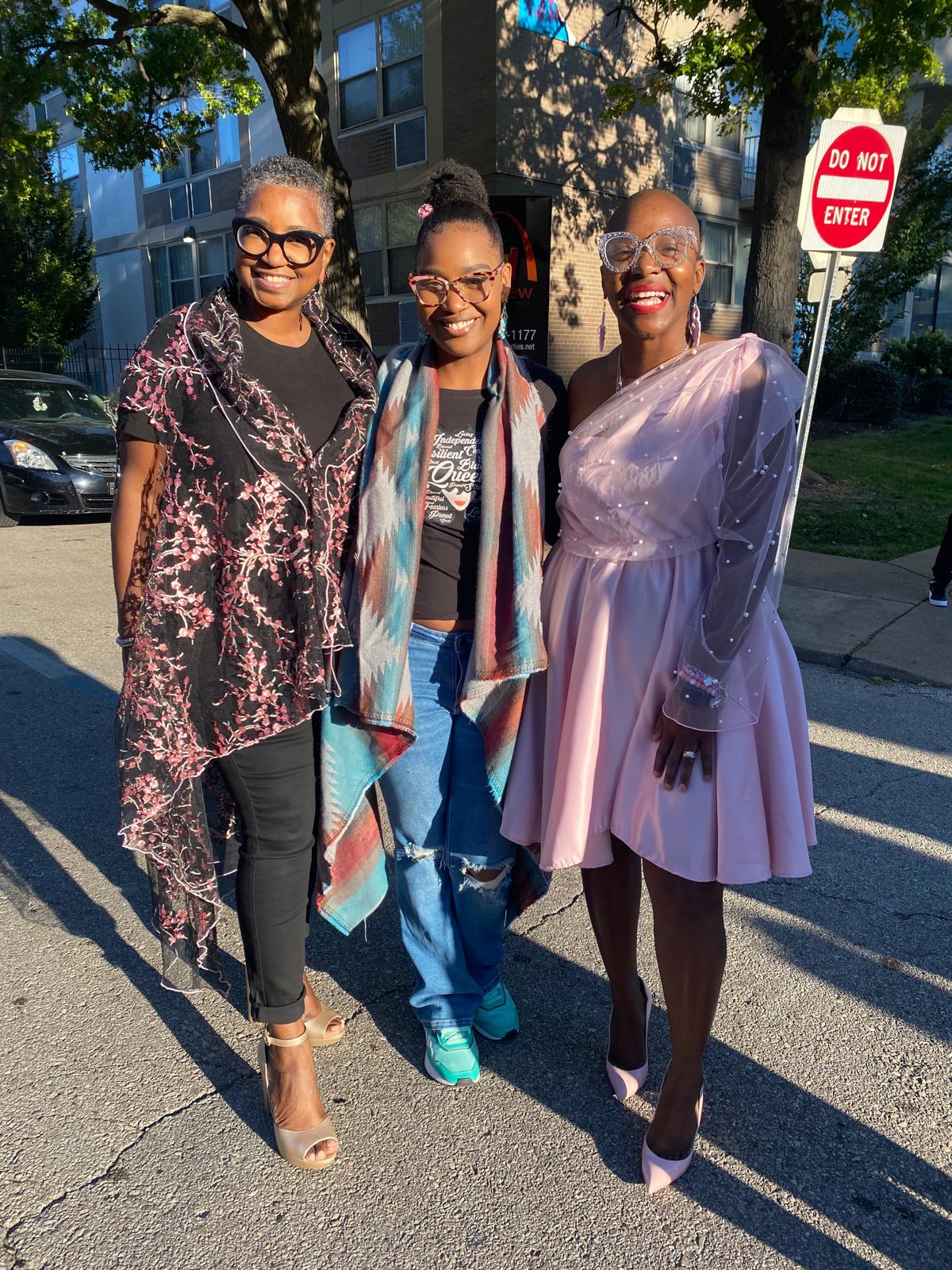 Three women standing outdoors near a crosswalk and a stop sign, smiling and posing for the photo.
