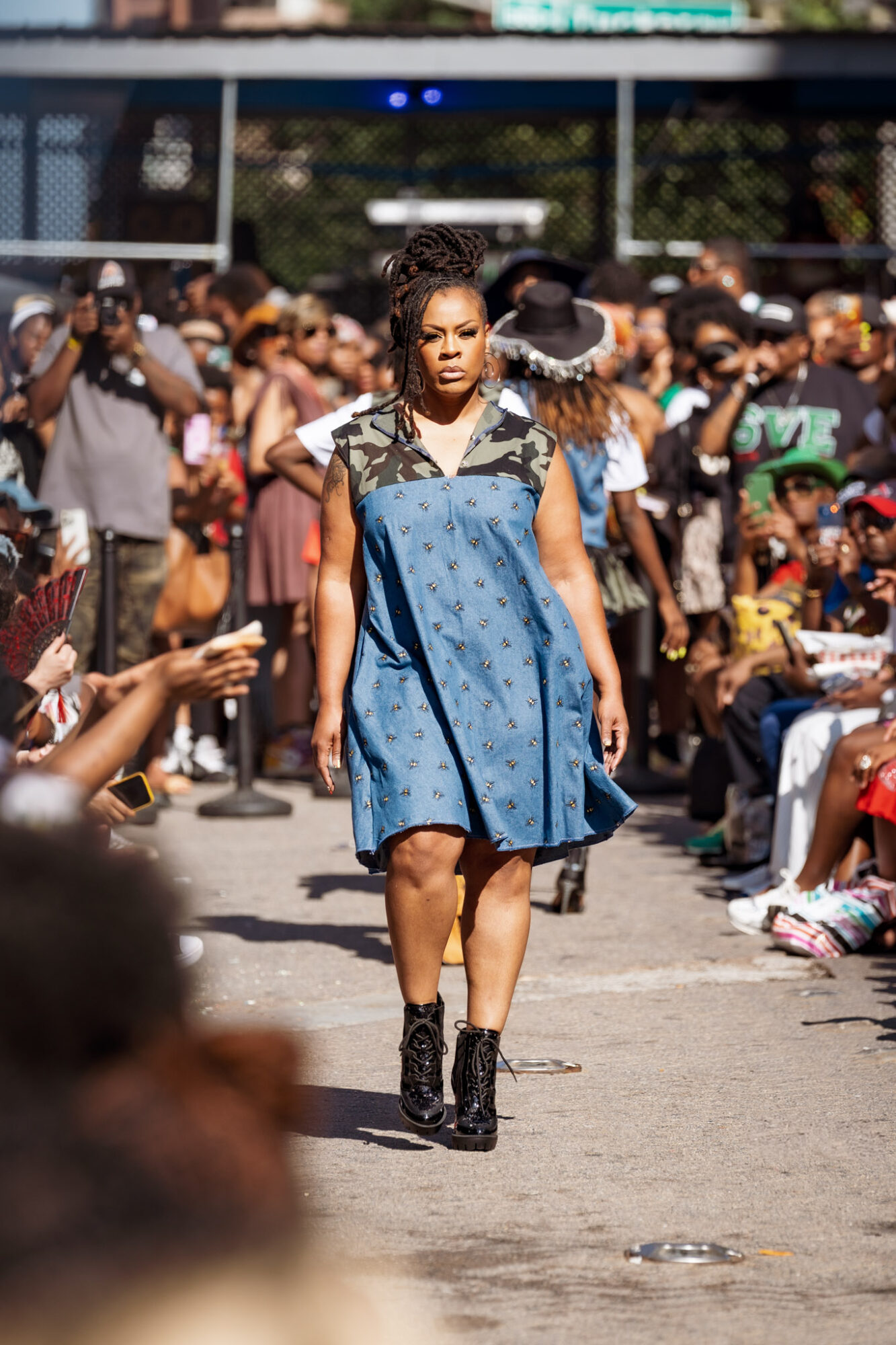 Young woman walking confidently in a crowd, wearing a blue dress and black boots, with people sitting and standing around.