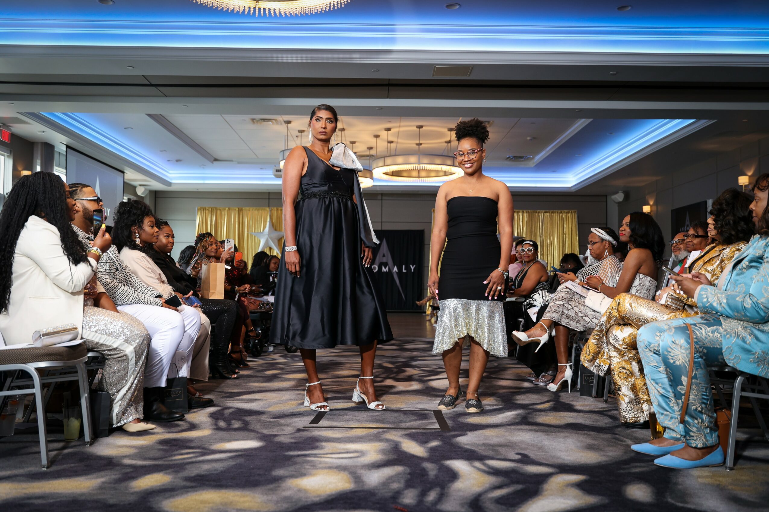 Two women walk down a runway in a fashion show, surrounded by seated audience members in a decorated room.
