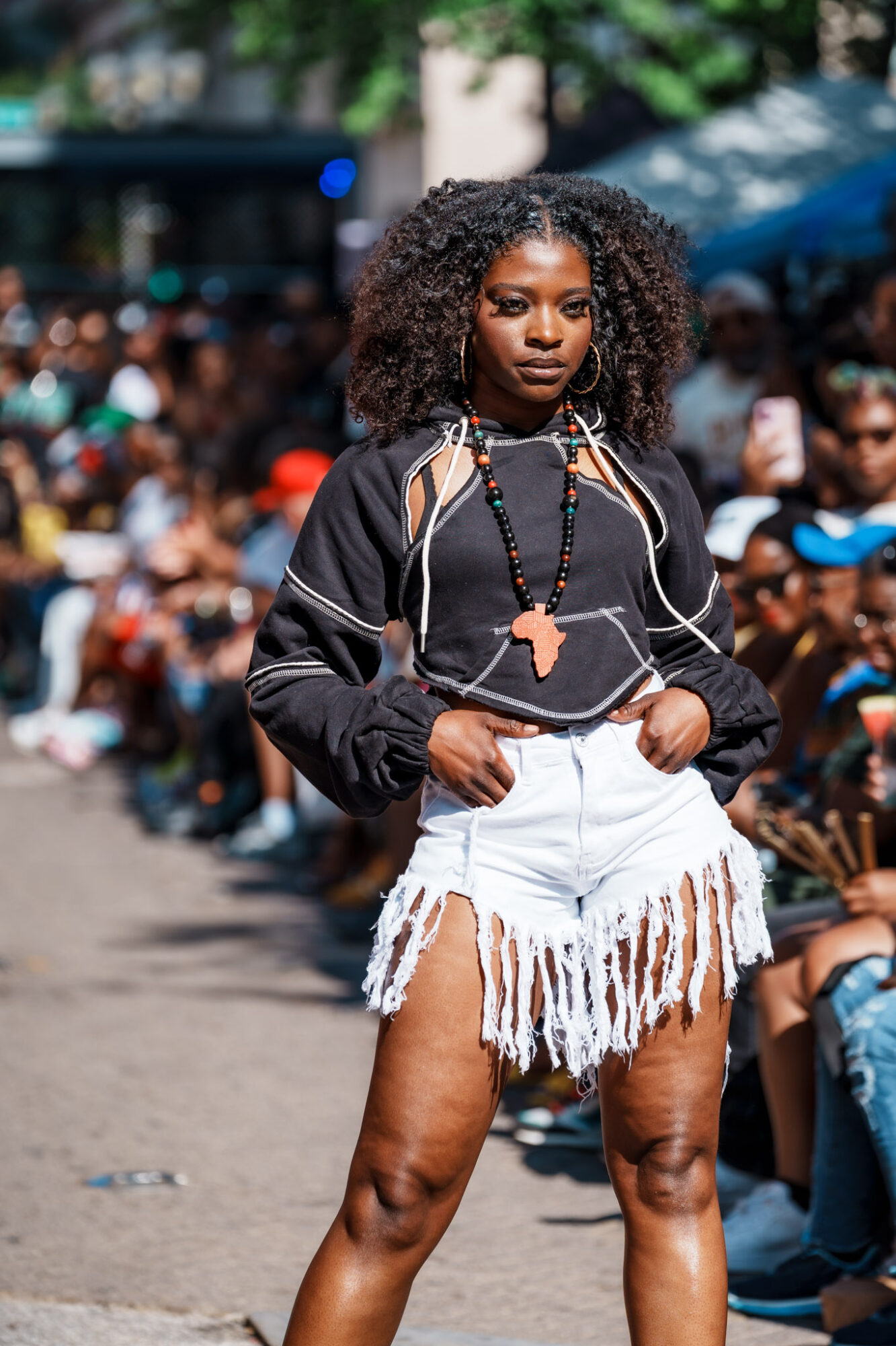 Young woman with curly hair stands confidently in a crowd outdoors, wearing a black top and white fringed shorts.