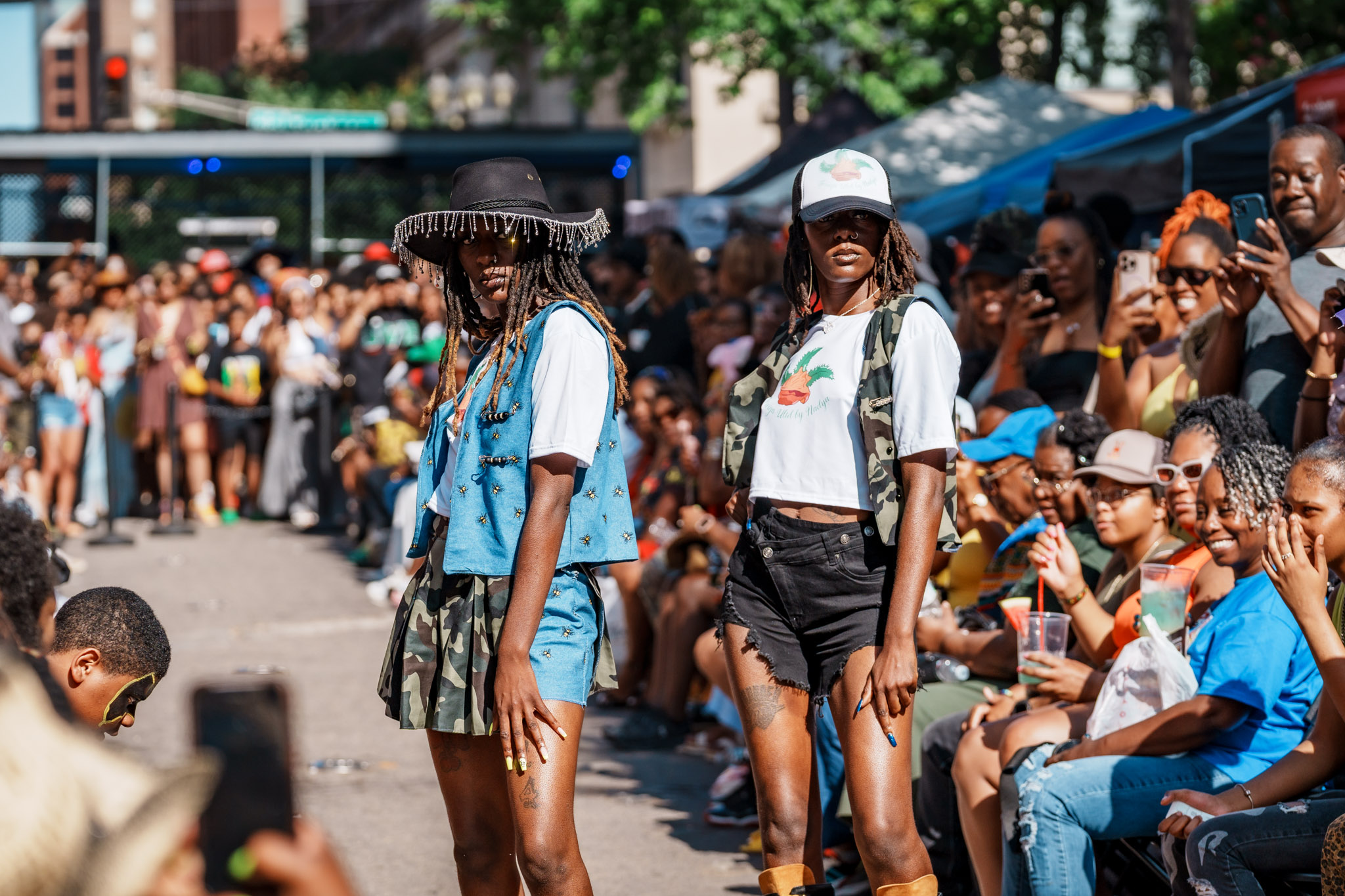 Two women standing in front of a large crowd at an outdoor event, with people sitting and standing around.