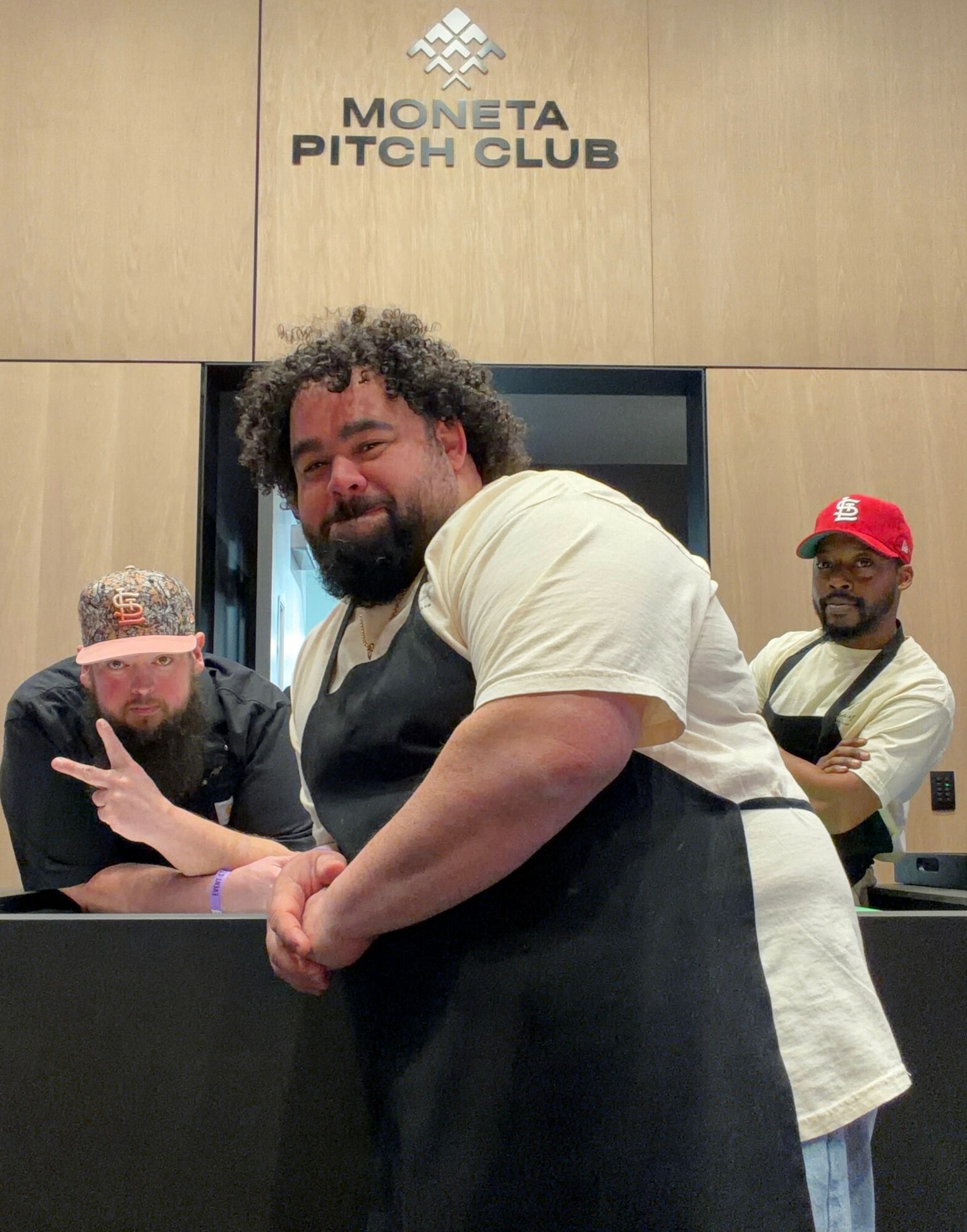 Three men standing in front of a wall with a sign that reads 'Moneta Pitch Club.' One man in the foreground has curly hair and a beard, wearing a white shirt and black apron. Two men in the background wear glasses and hats, one with a red cap and the other with a patterned cap.
