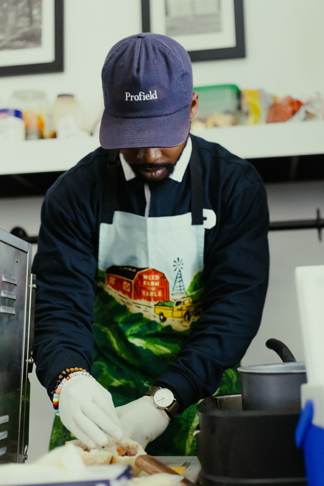 Person wearing a blue cap, apron, and gloves preparing food in a kitchen, with shelves and containers in the background.