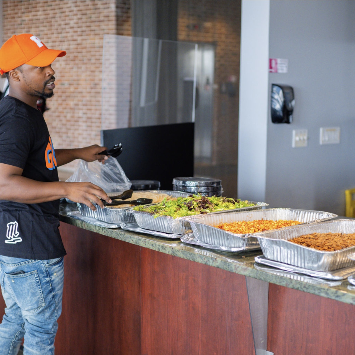 Person in black shirt and orange cap serving food at a buffet table with trays of dishes, in an indoor setting.