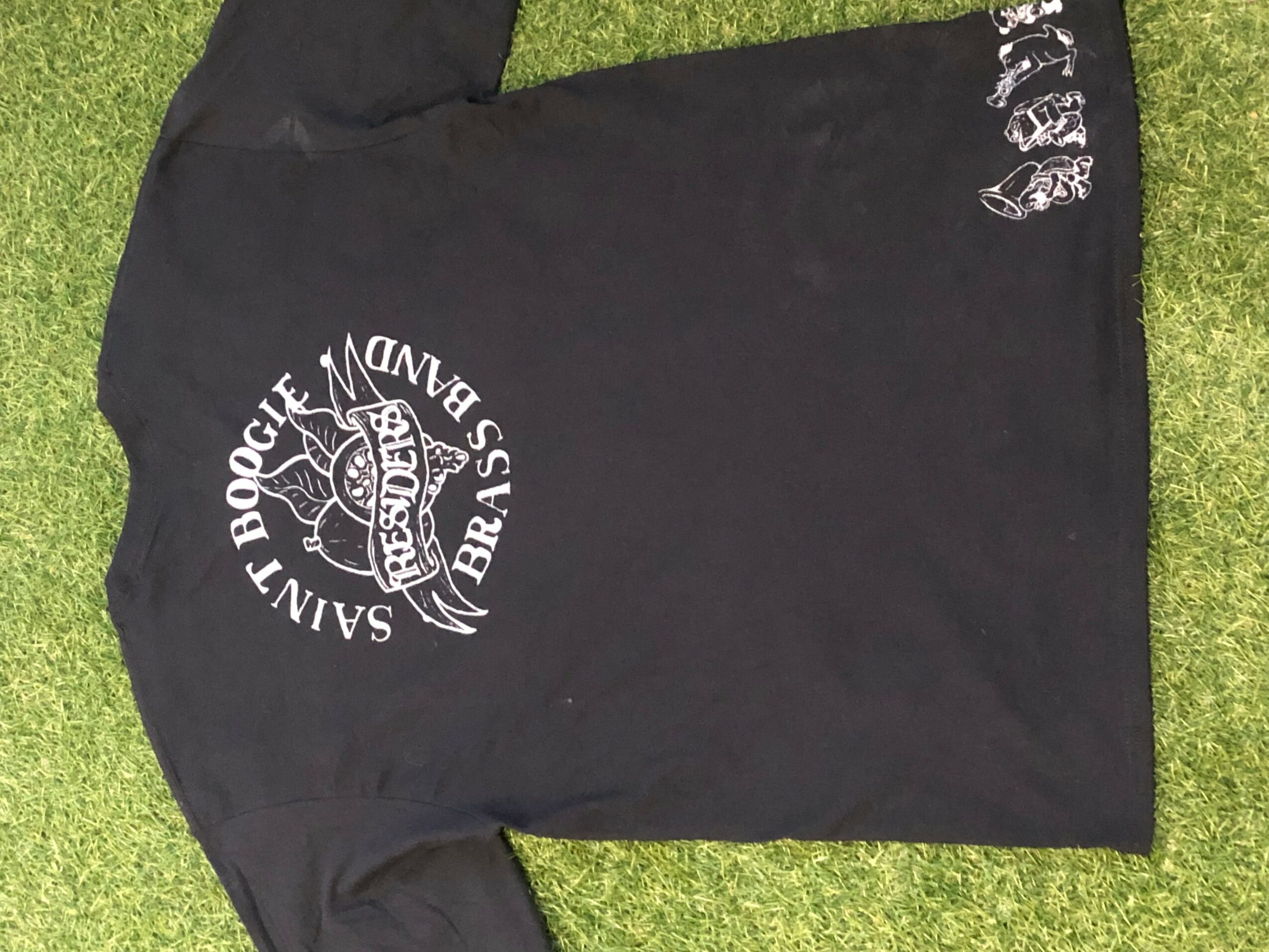 Black T-shirt with white logo and text, laid flat on grass, with some white text near the bottom edge.